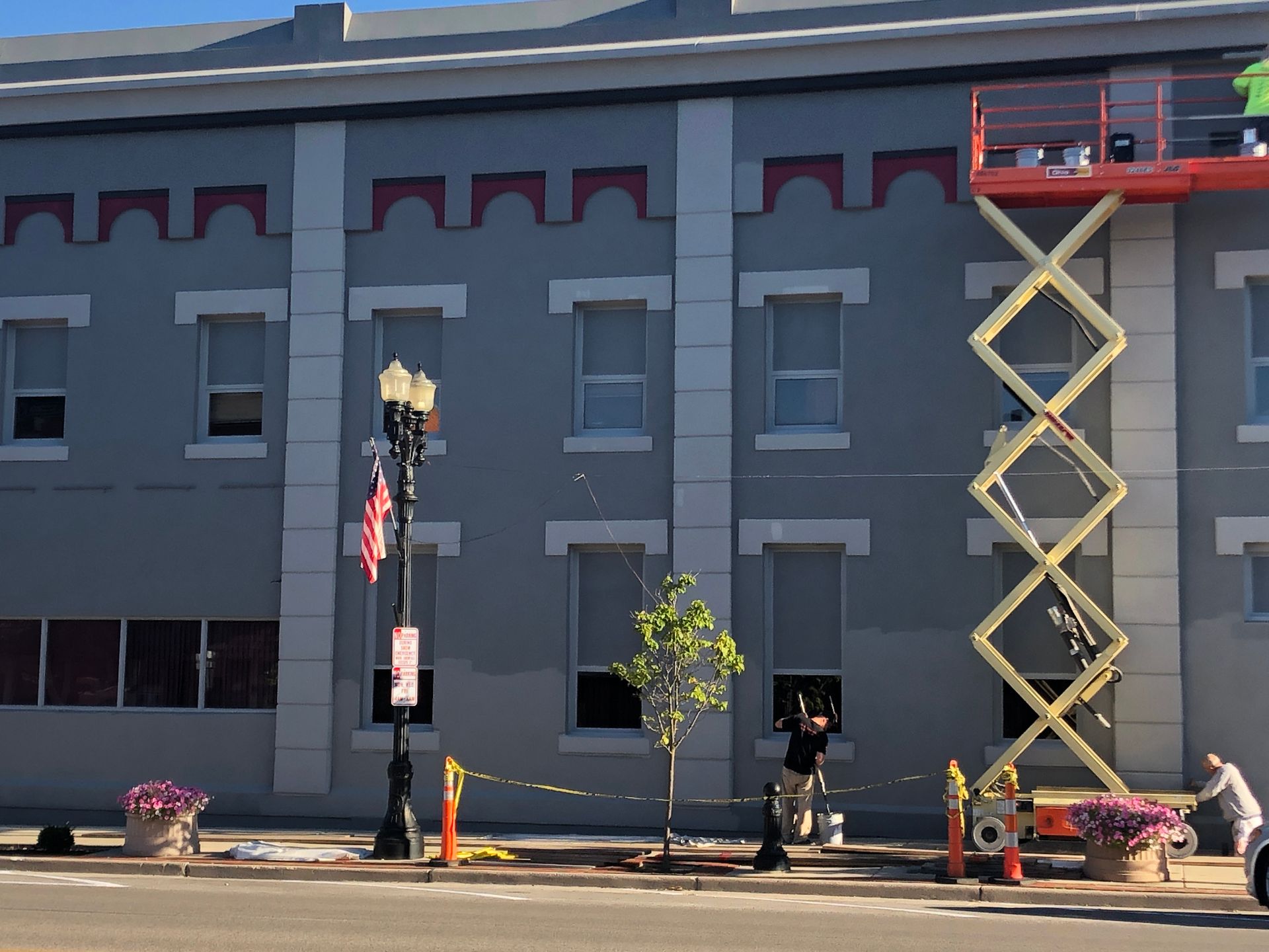 Gray building facade with a raised scissor lift and workers near a streetlight and potted plants