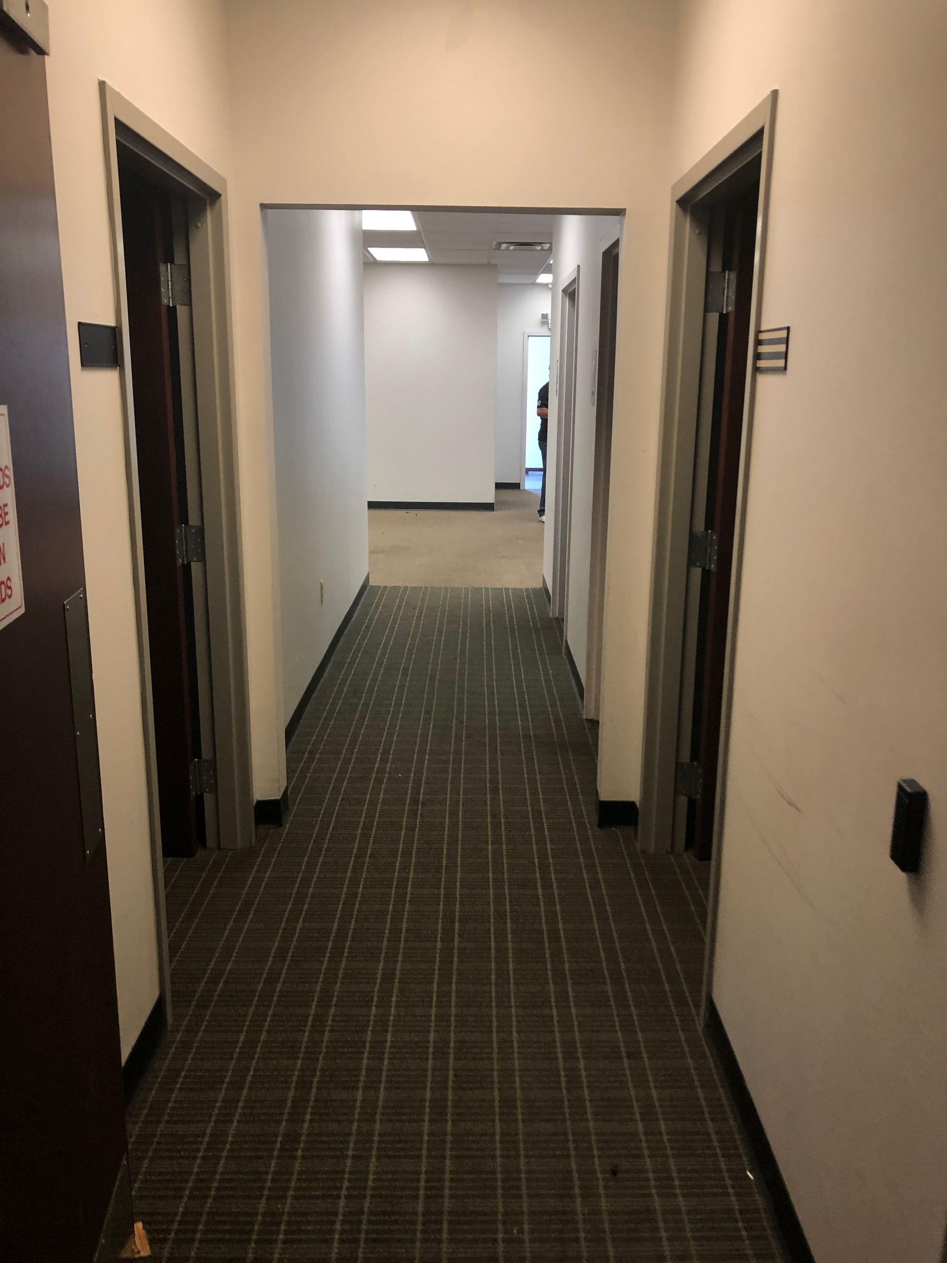 Narrow office hallway with dark carpet, beige walls, and open doorways on both sides