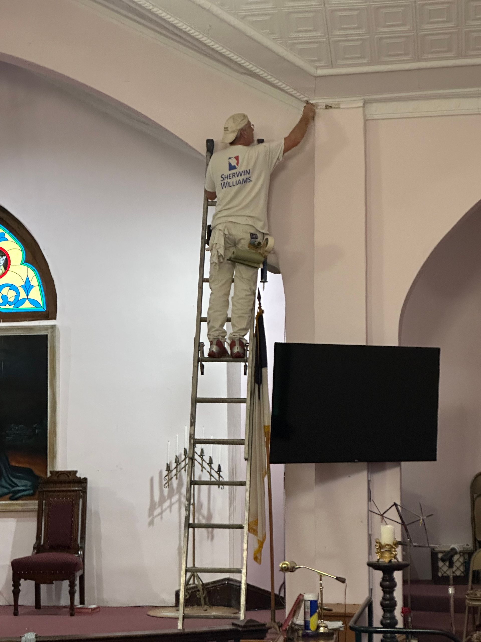 Worker on a ladder painting a church ceiling arch beside a wall-mounted TV