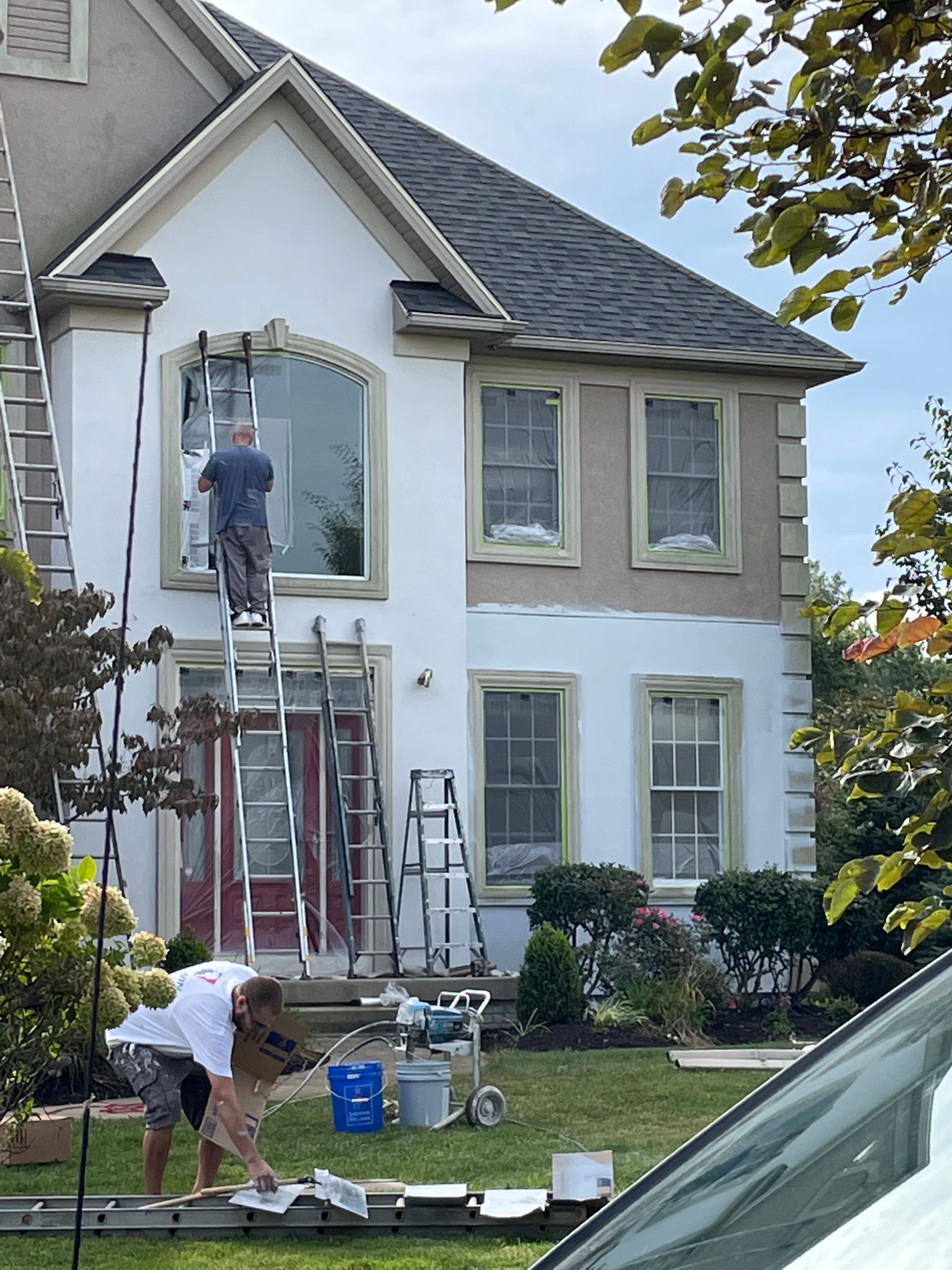 Two workers painting a house exterior beside ladders and supplies