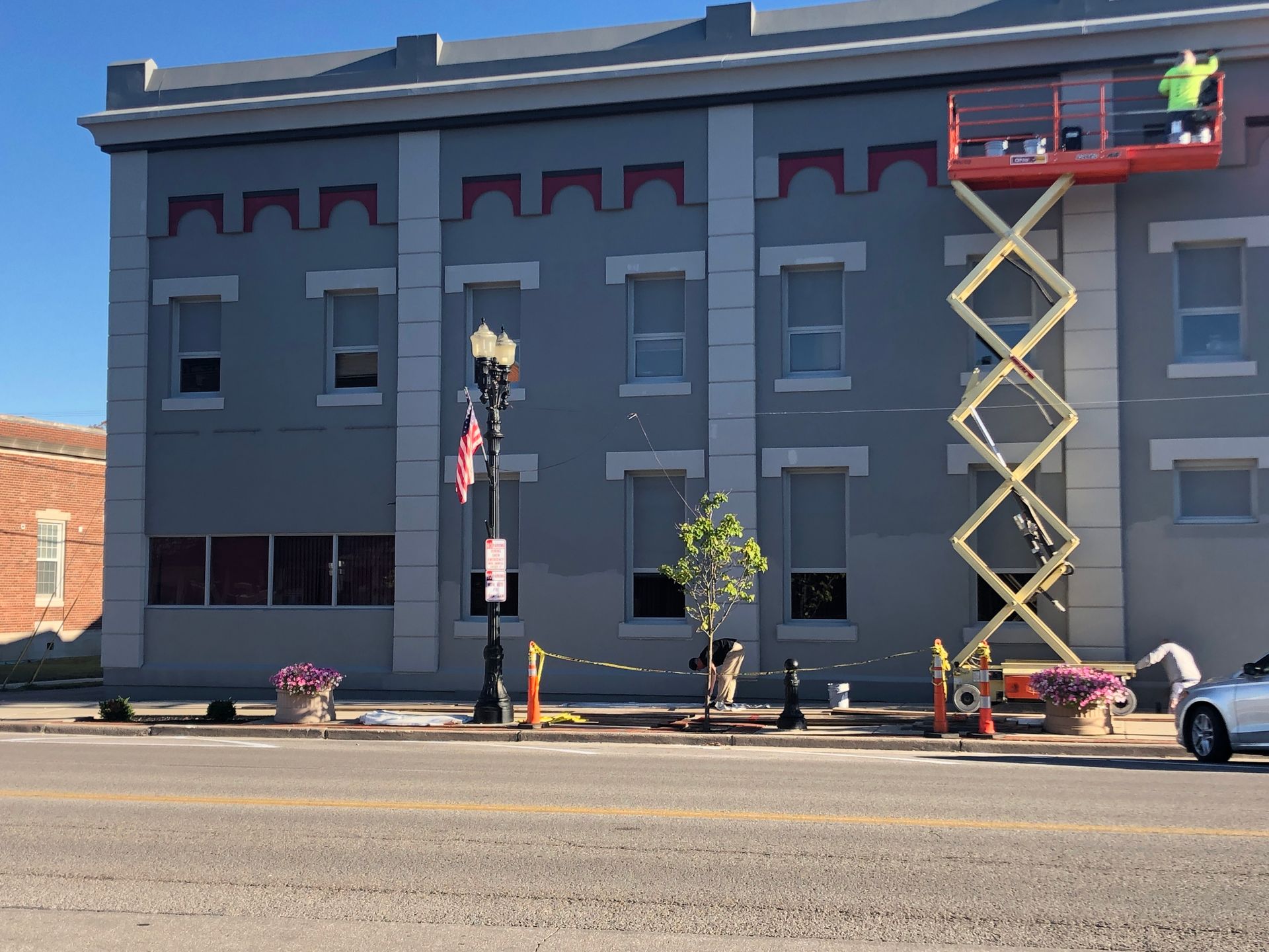 Gray building with workers on a scissor lift and a cherry picker outside on a city street