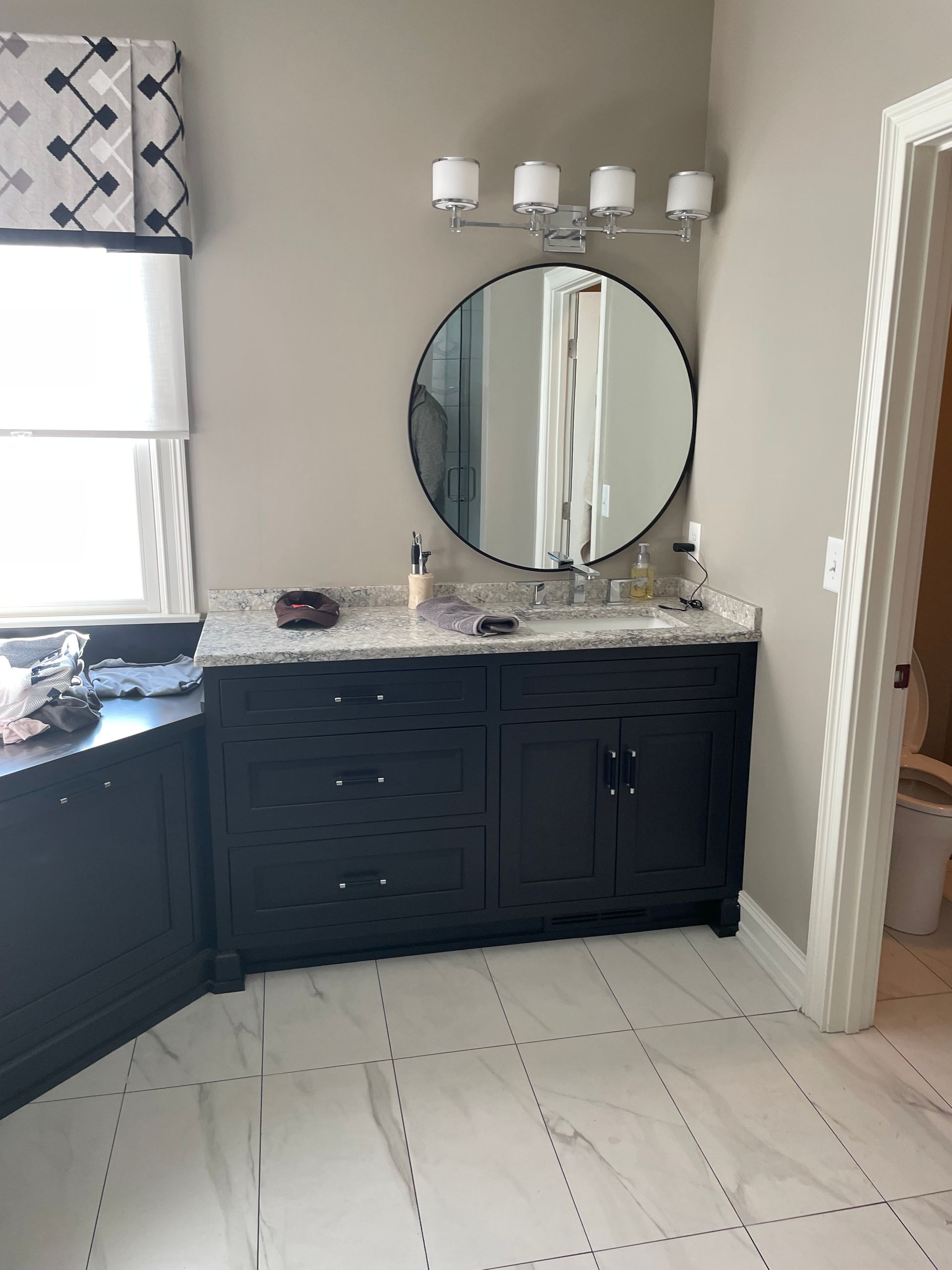 Bathroom vanity with dark cabinets, round mirror, and white tile floor near a window.