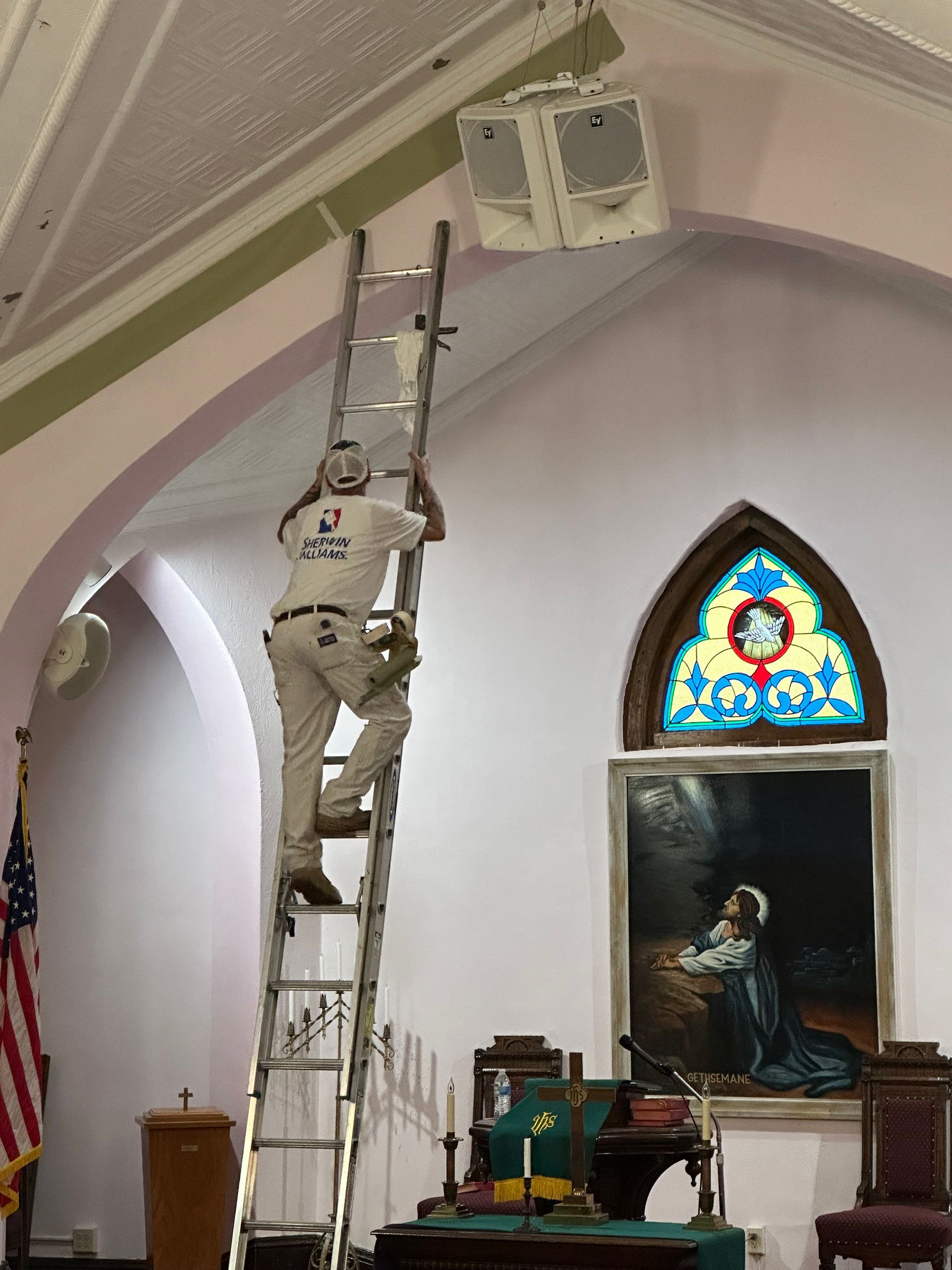 Worker on a ladder adjusting a ceiling-mounted fixture inside a church sanctuary.
