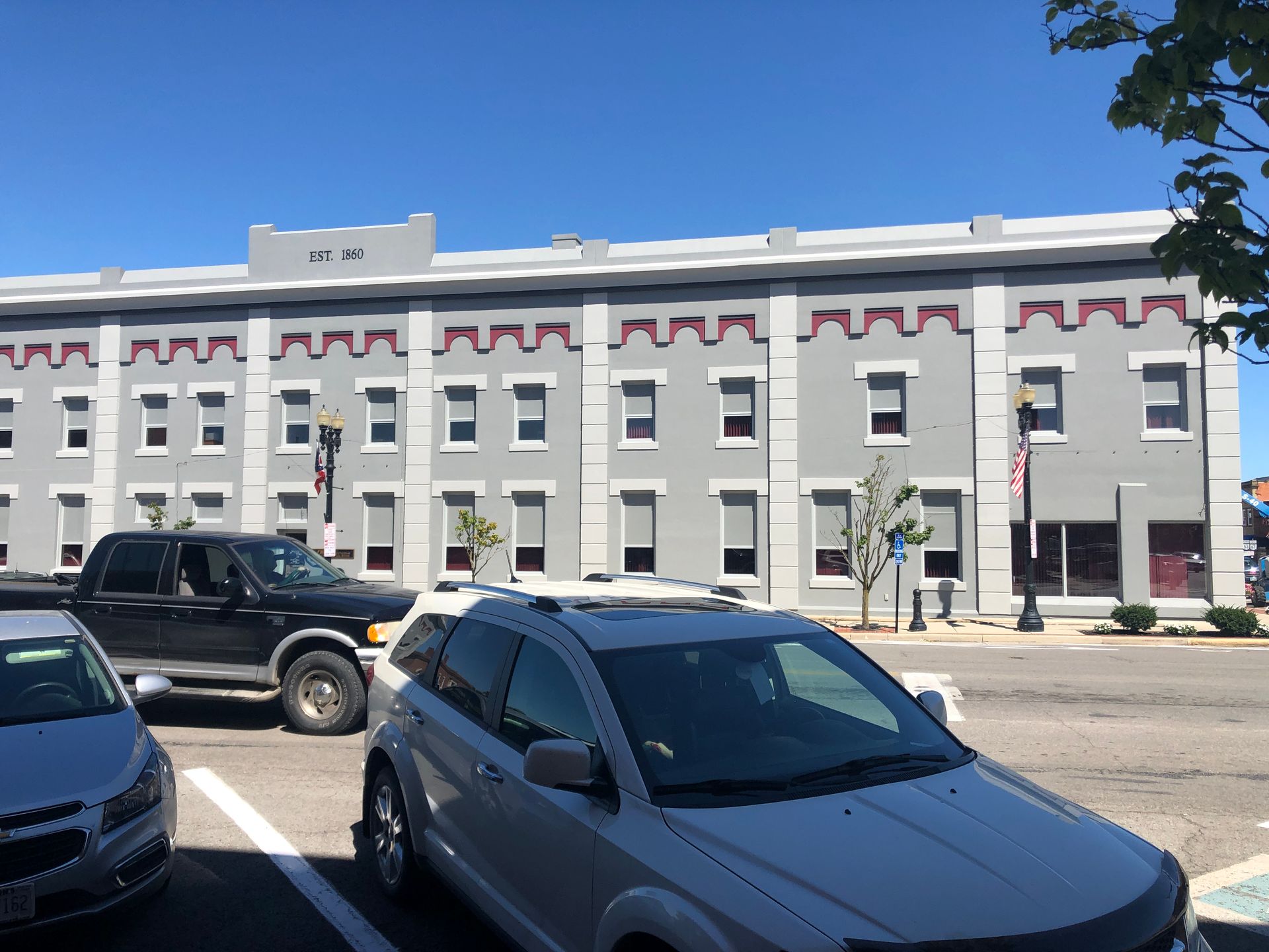 White historic building with red trim on a sunny street, with parked cars in front.