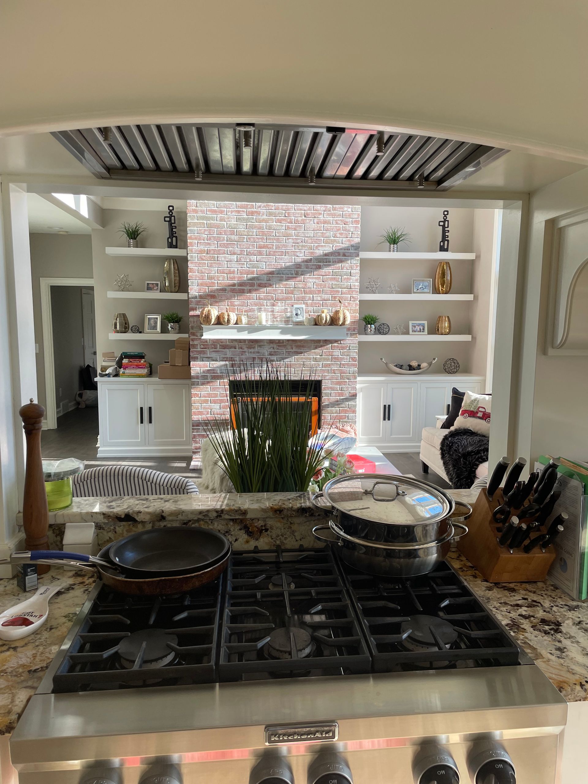 Kitchen view from stovetop, with two white shelves and a brick backsplash above a gas range.