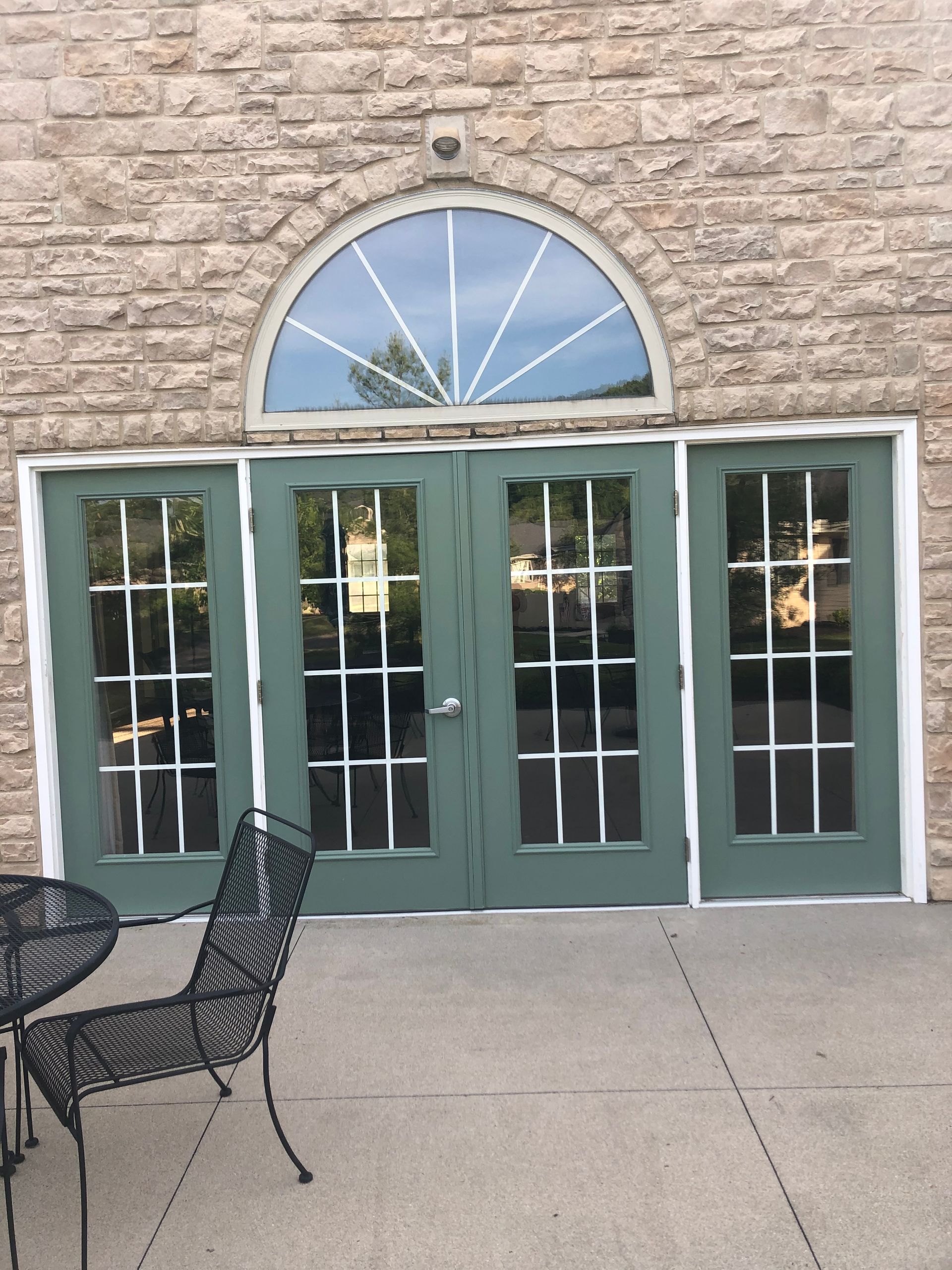Stone building entrance with green double doors and arched window, beside an outdoor patio with black chairs