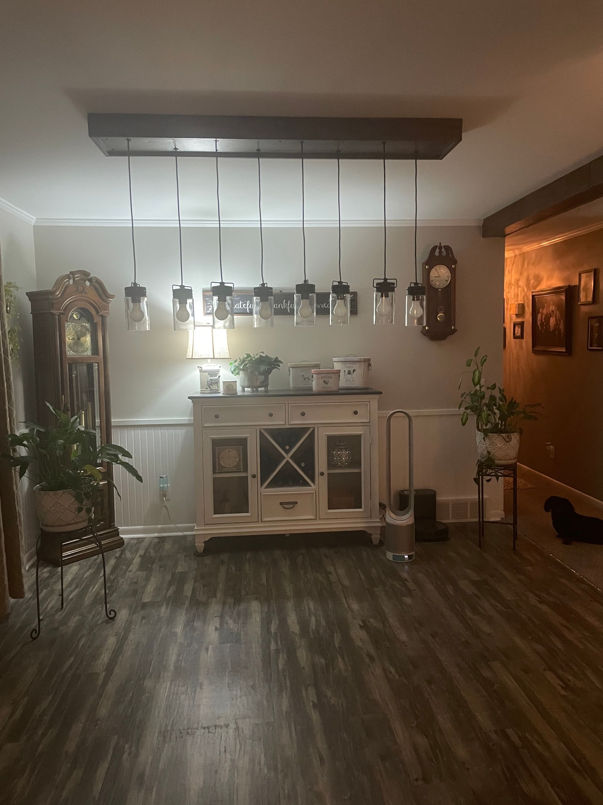 Cozy dining room with hanging lights, a white sideboard, wall clock, plants, and wood flooring