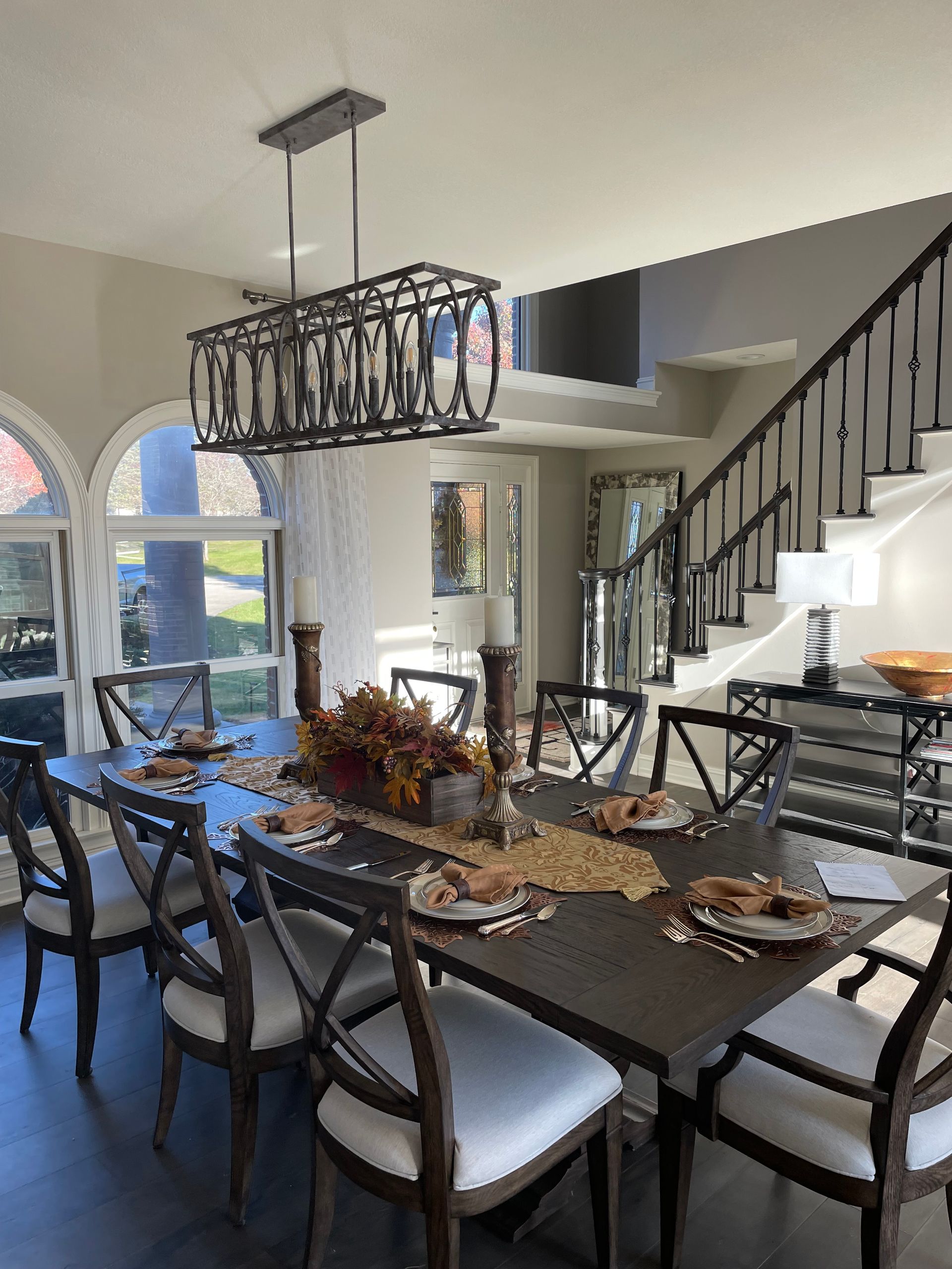 Dining room with set table, dark chairs, chandelier, and staircase beside large windows.