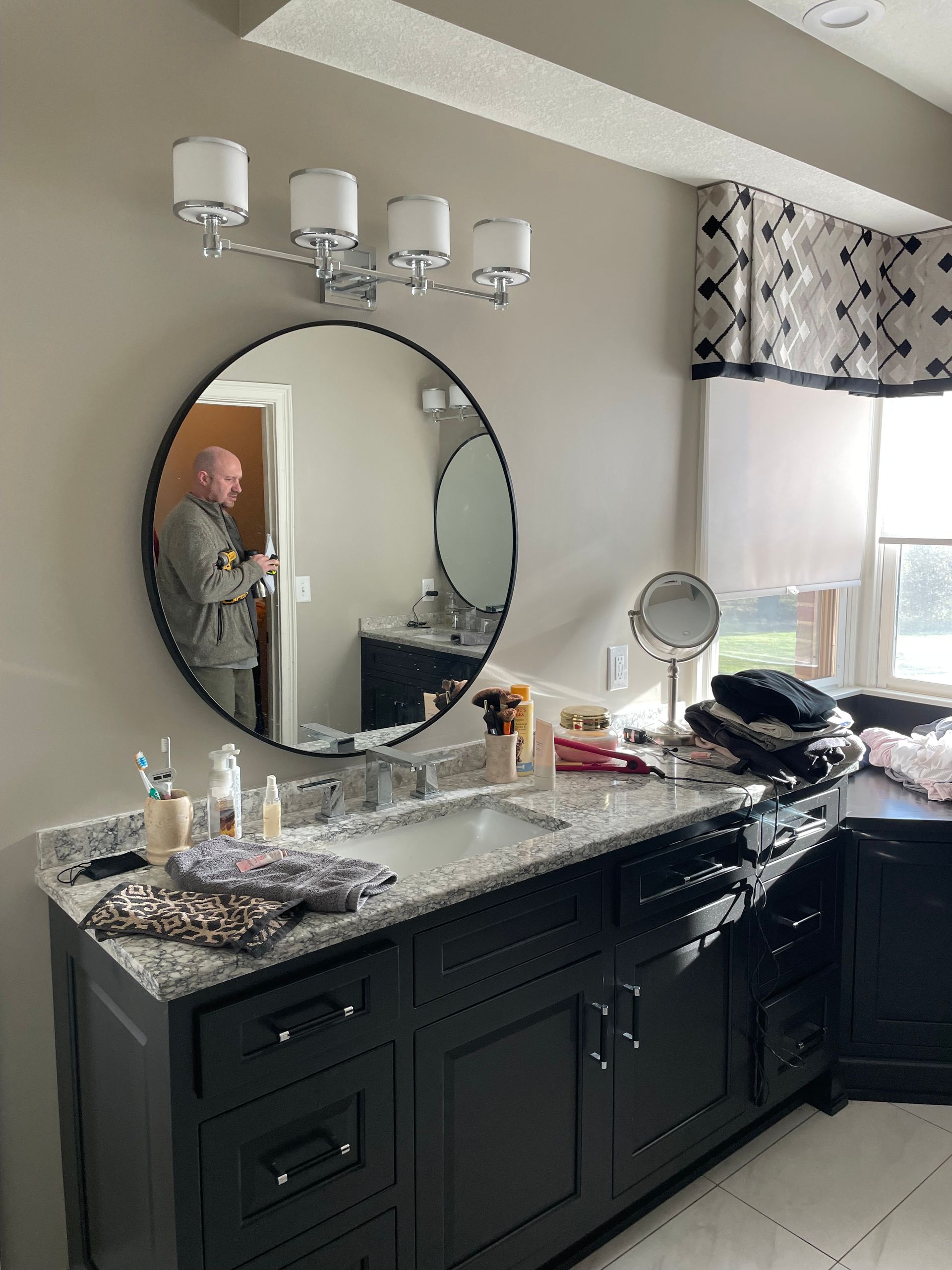 Bathroom vanity with round mirror, granite counter, and black cabinets; person visible in mirror reflection