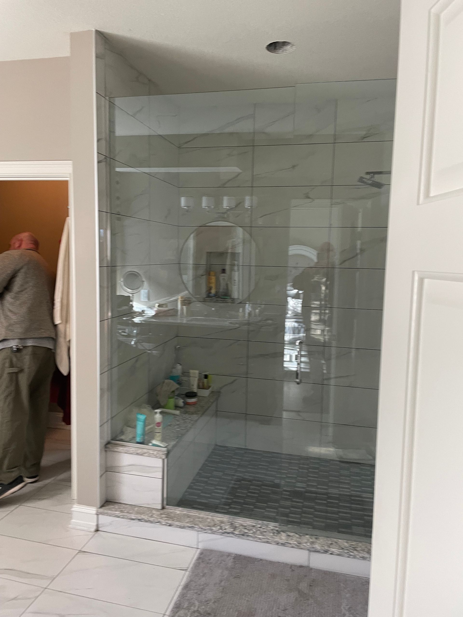 Glass shower enclosure in a bathroom with tiled floor and walls; person partially visible in adjacent room.