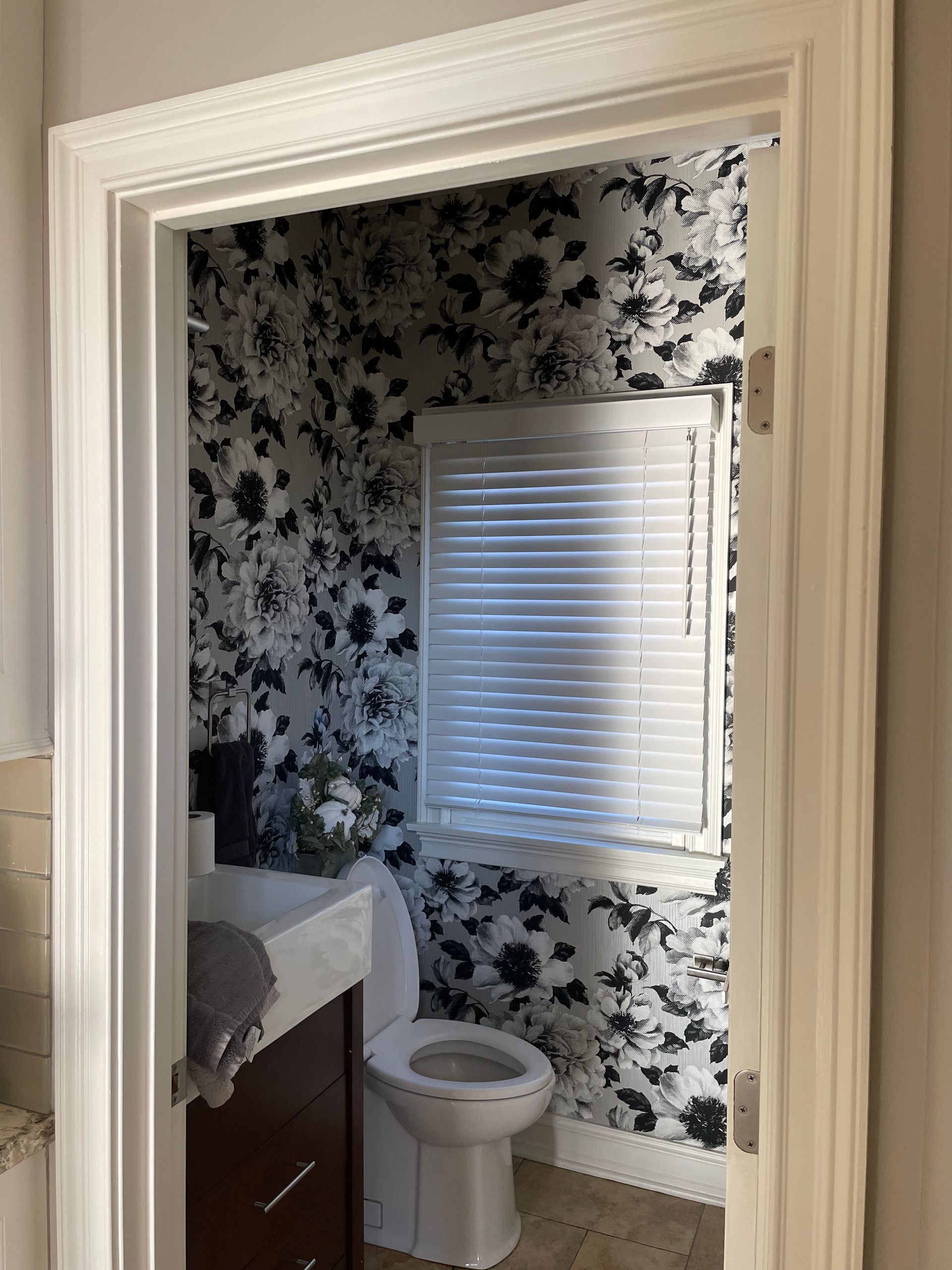 Small bathroom with black-and-white floral wallpaper, a window, toilet, and sink vanity visible through a doorway