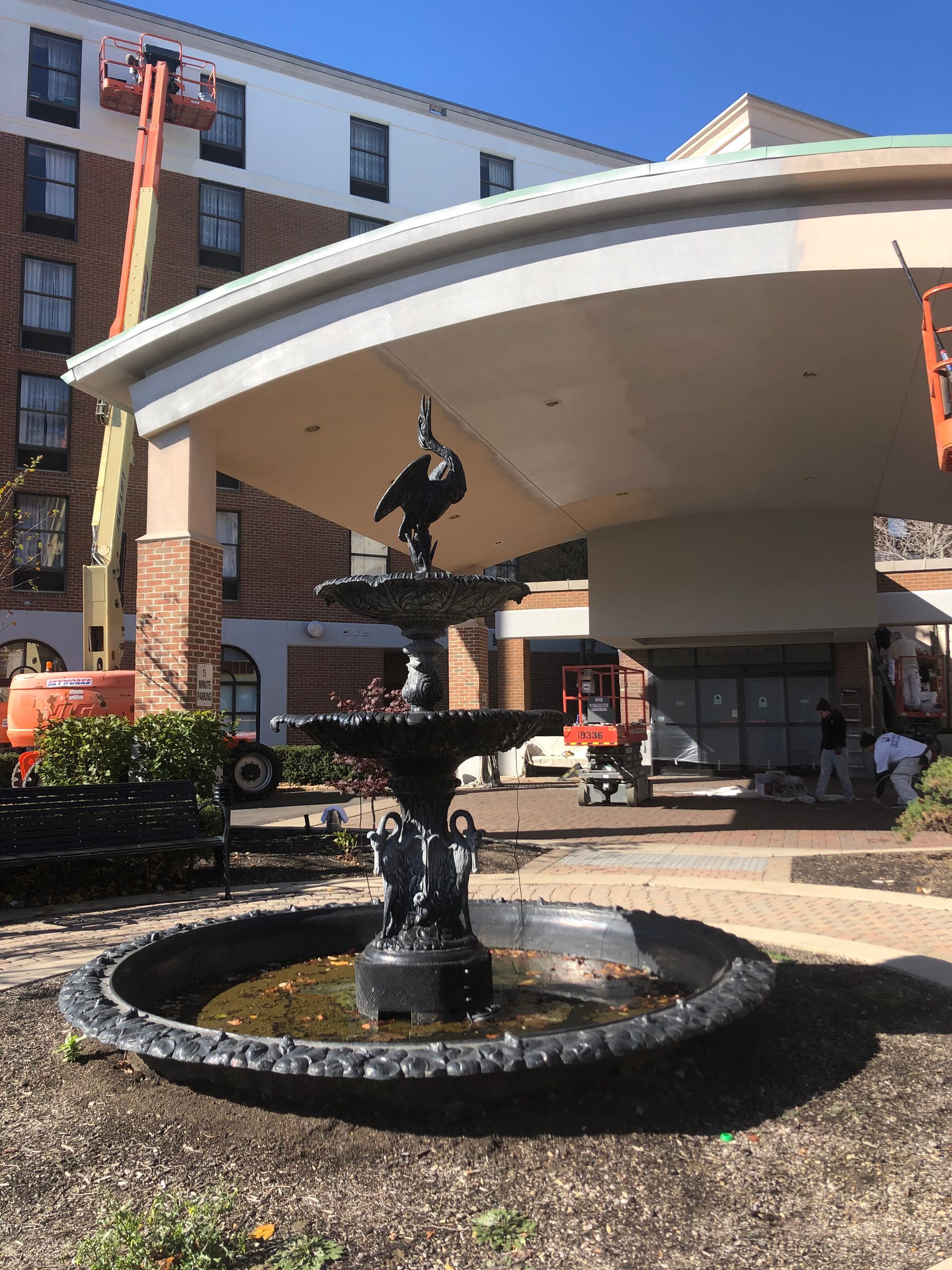 Courtyard fountain under a large curved canopy beside a brick building, with hanging sculpture and outdoor seating.