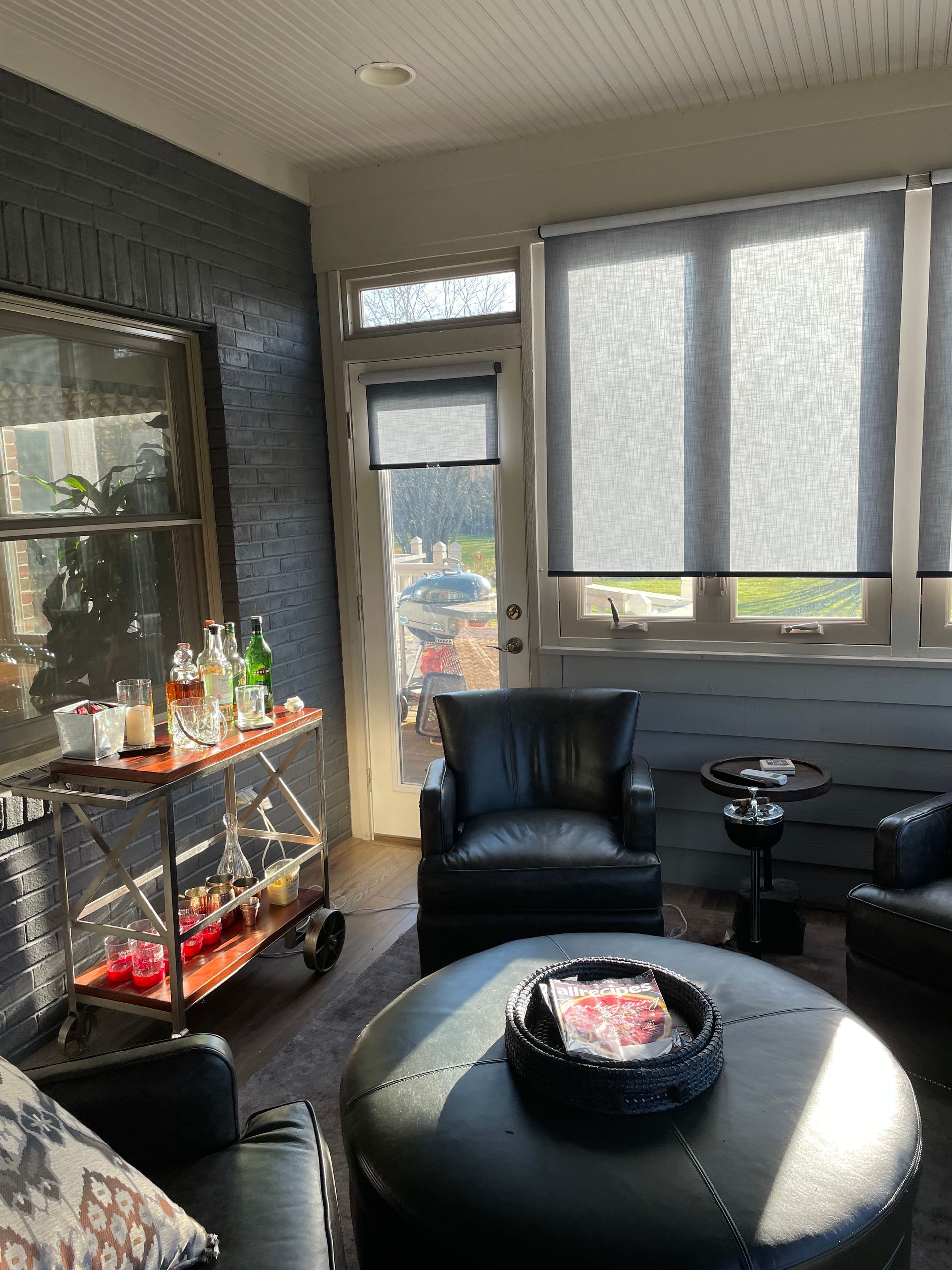 Sunlit screened porch with black chair, glass table, and small bar cart by windows