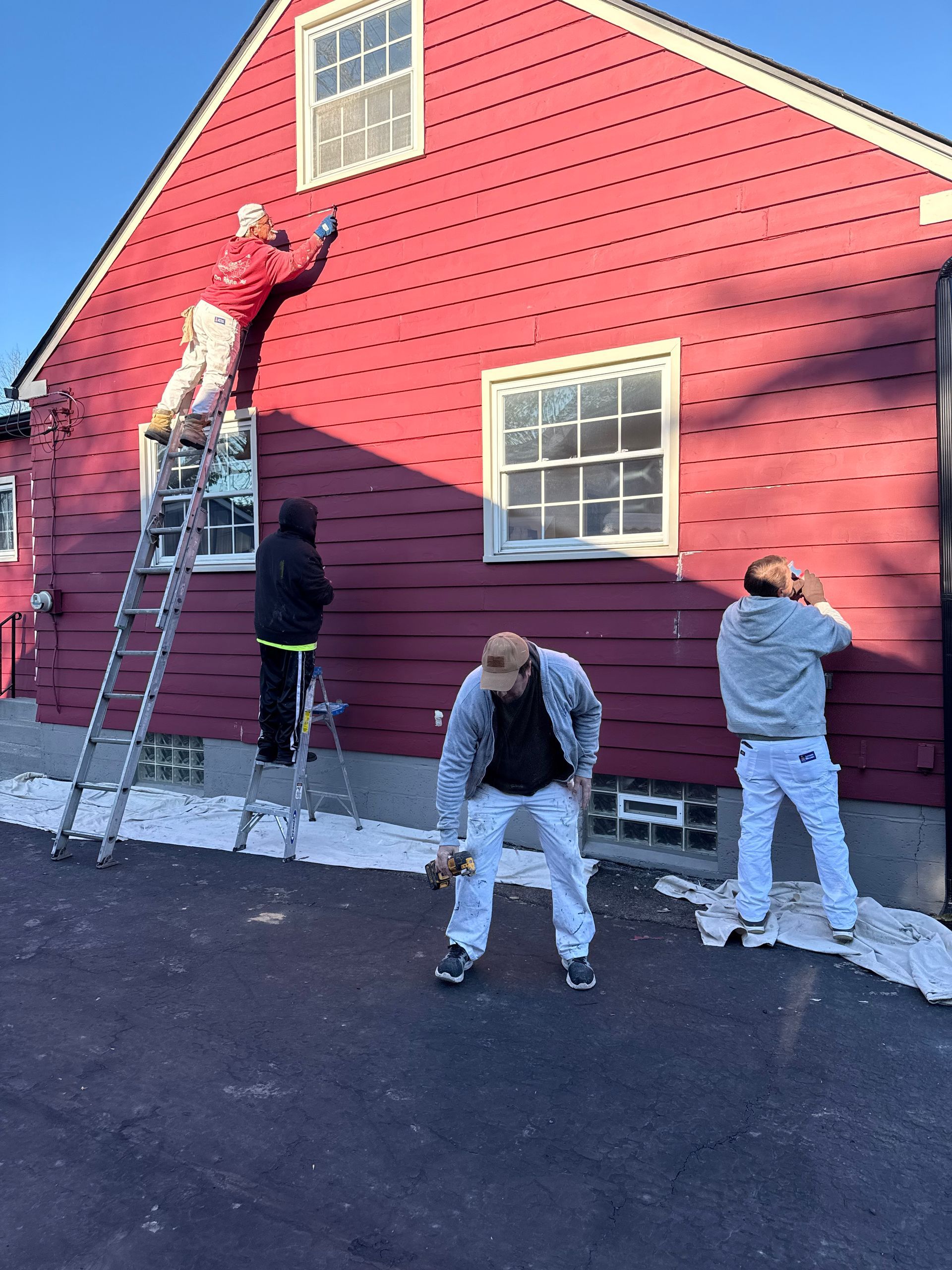 People painting a red house exterior, with one on a ladder and others working below.