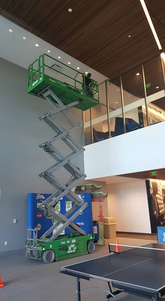 A man is standing on a scissor lift in a building next to a ping pong table.