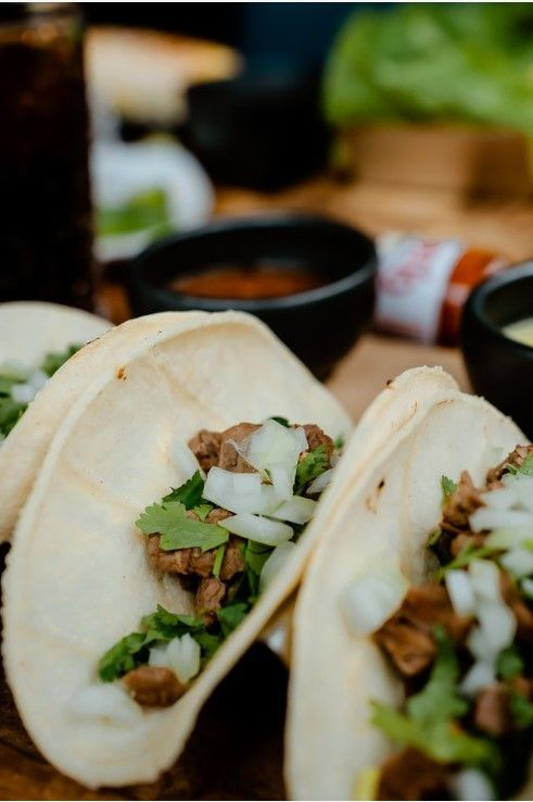 Three tacos are sitting on a wooden cutting board next to two bowls of sauce.