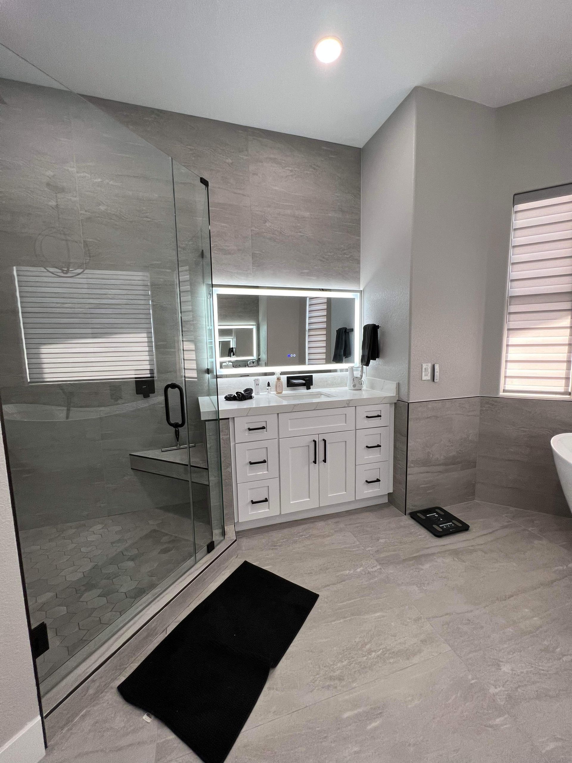 Modern bathroom with gray tiled walls, glass shower, white vanity, and black bath mat.
