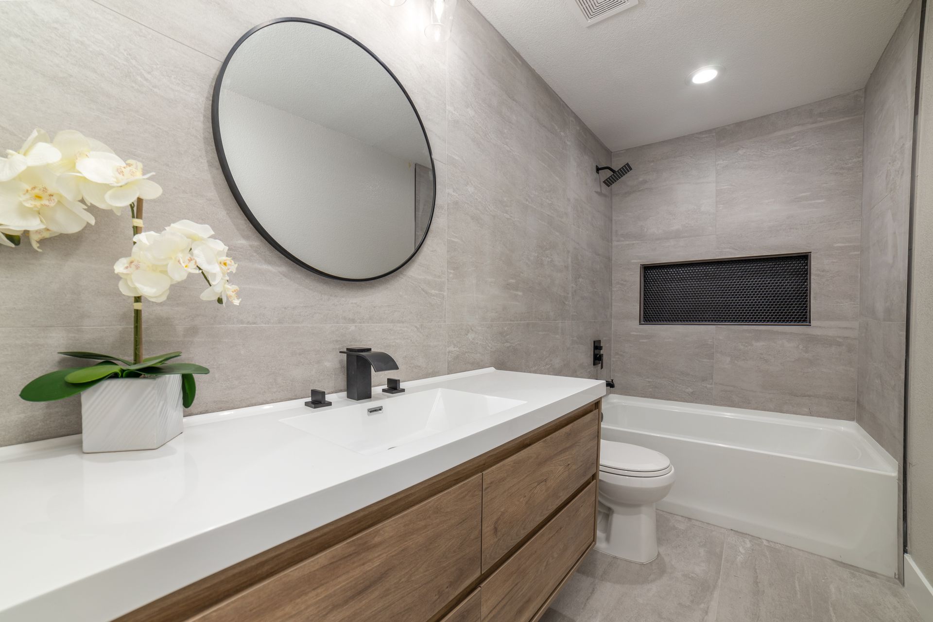 Modern bathroom with gray tiled walls, round mirror, white sink, wooden vanity, and white orchid.