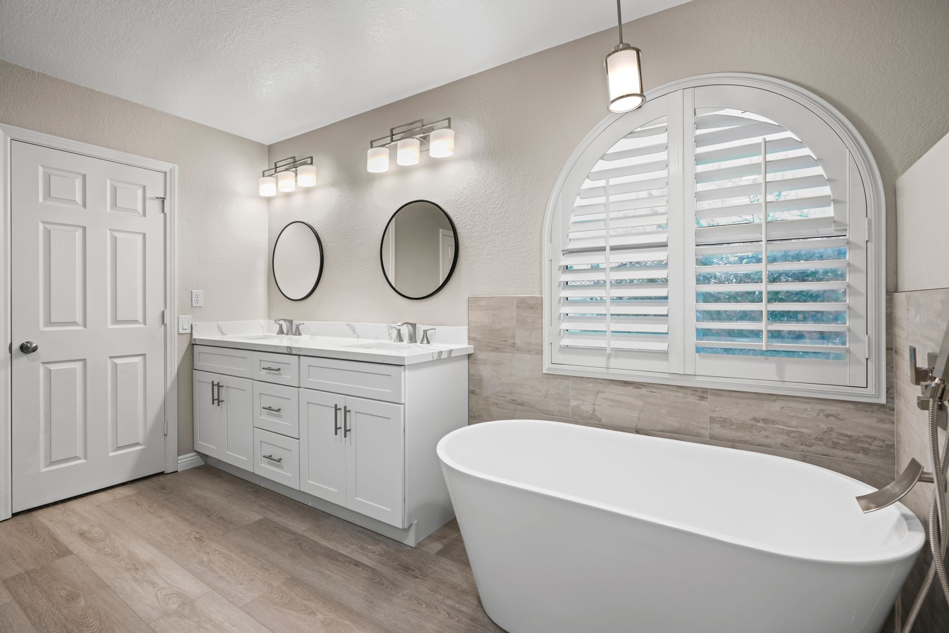 White bathroom with double vanity, oval tub, arched window with shutters.