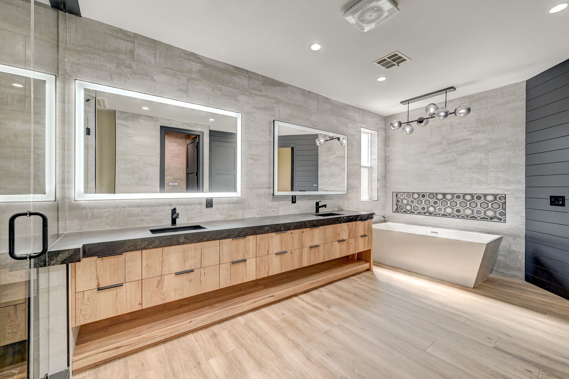 Modern bathroom with light wood cabinets, gray tiled walls, two illuminated mirrors, and a freestanding tub.