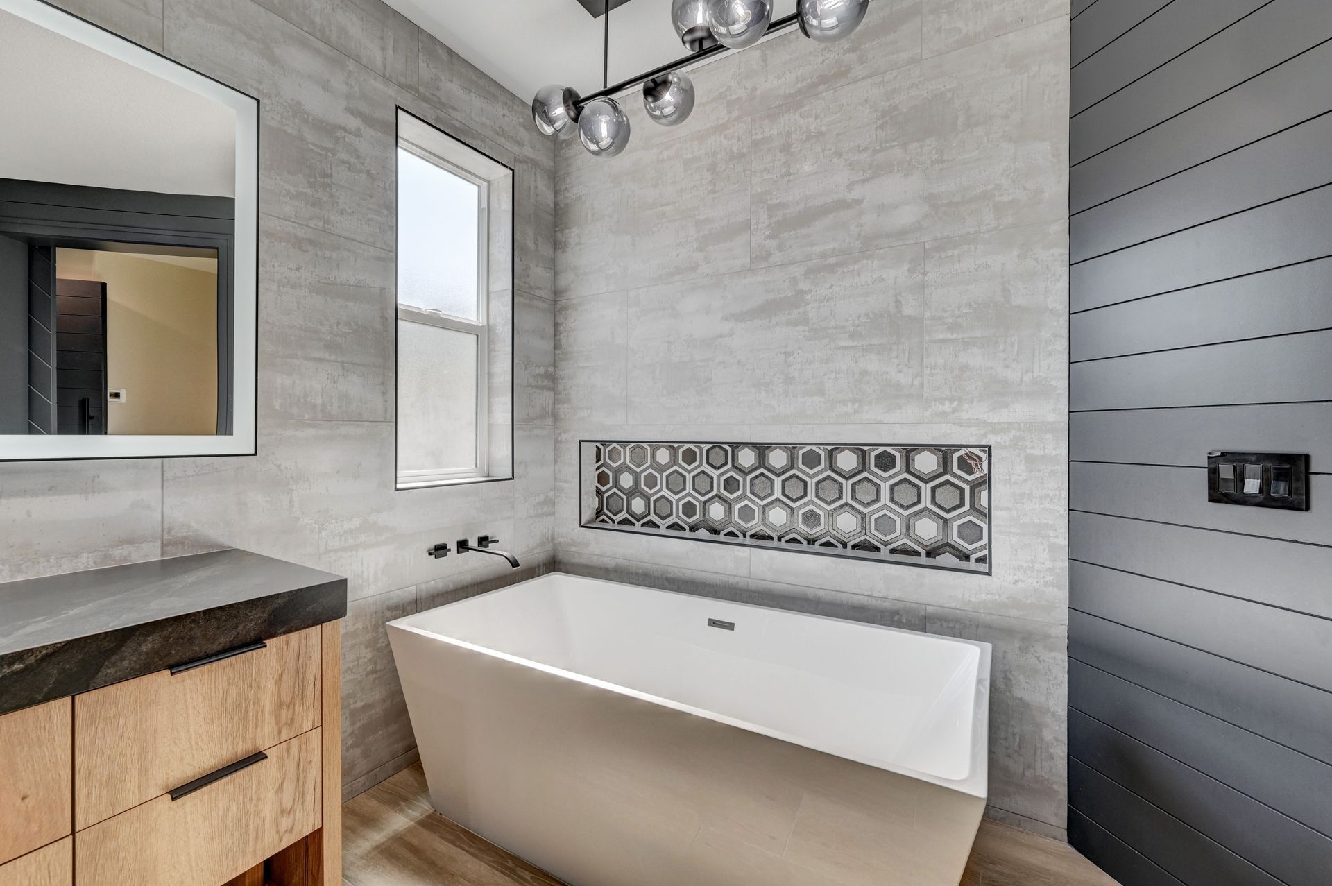 Modern bathroom with a white tub, gray tiled walls, and wood vanity.
