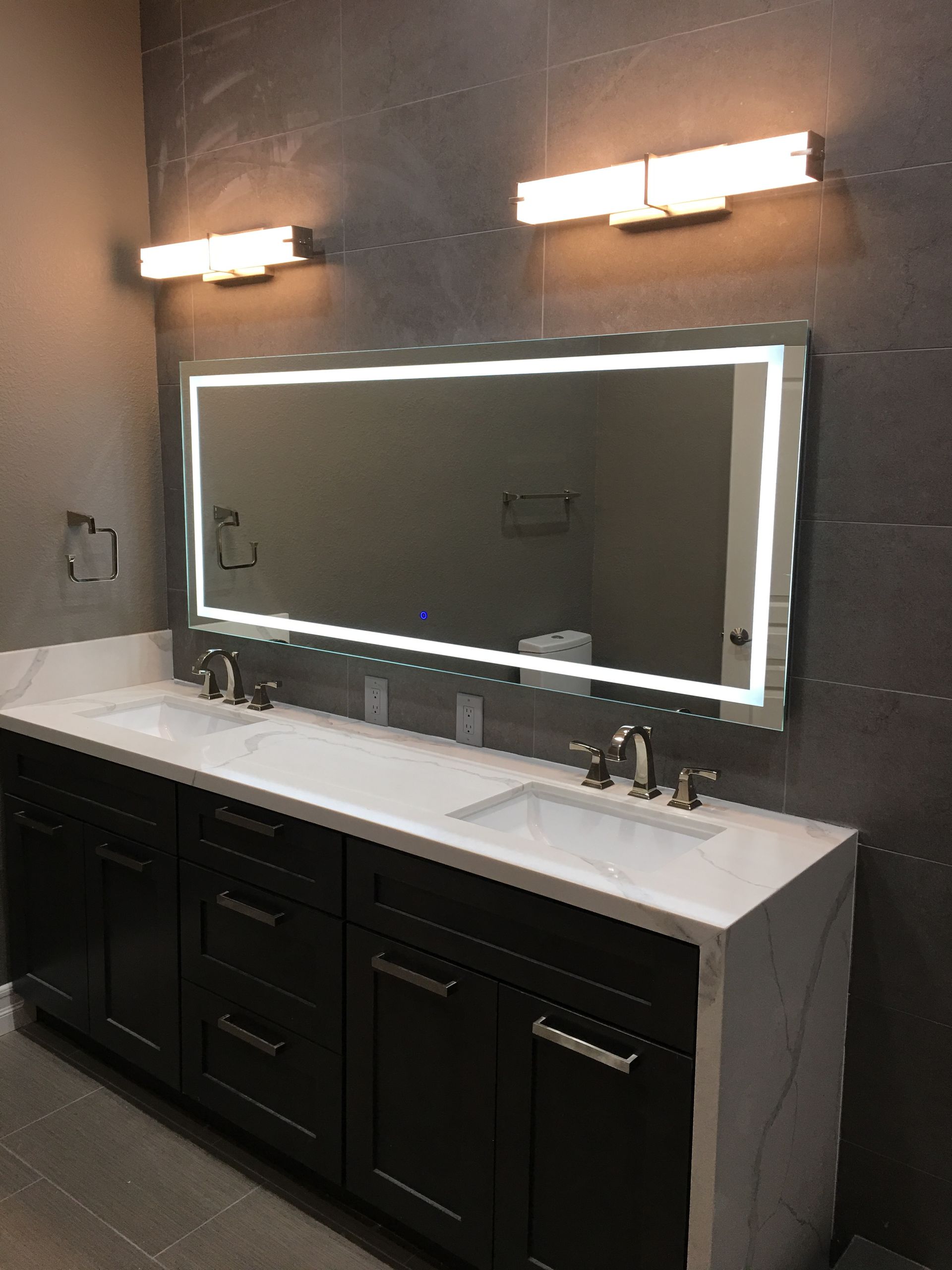Bathroom with dark wood vanity, white countertop, illuminated mirror, two sconces, and gray tiled wall.