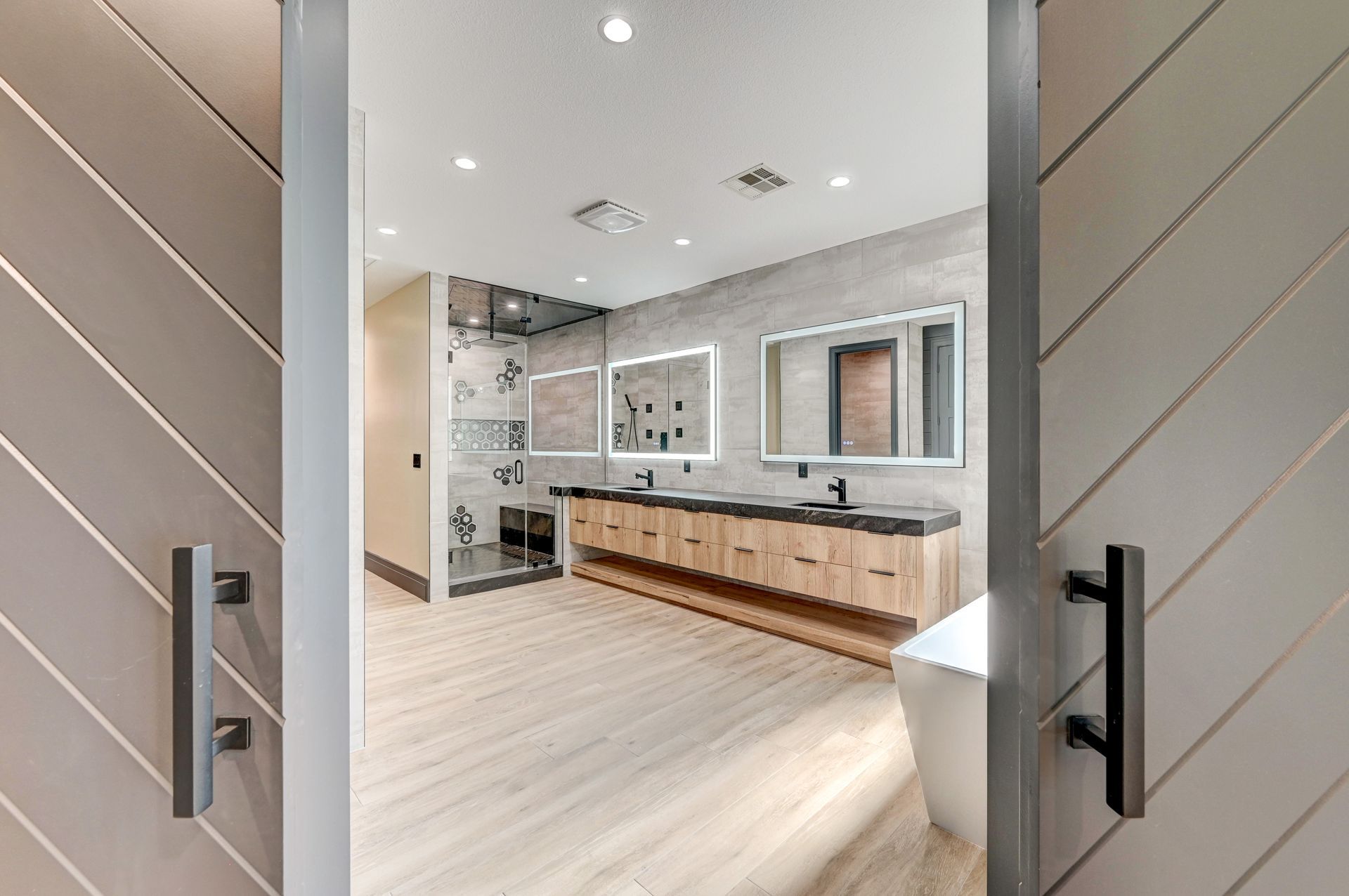 Bathroom interior, viewed through open sliding doors. Light wood floors, wood vanity, rectangular mirrors, glass shower, and freestanding tub.