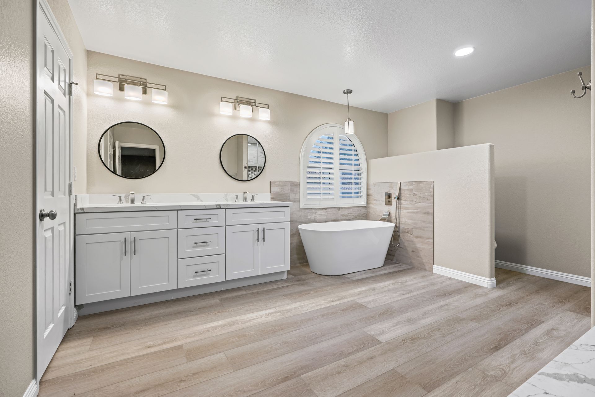Spacious, modern bathroom with double vanity, two round mirrors, and a freestanding tub. Light wood floors and neutral walls.