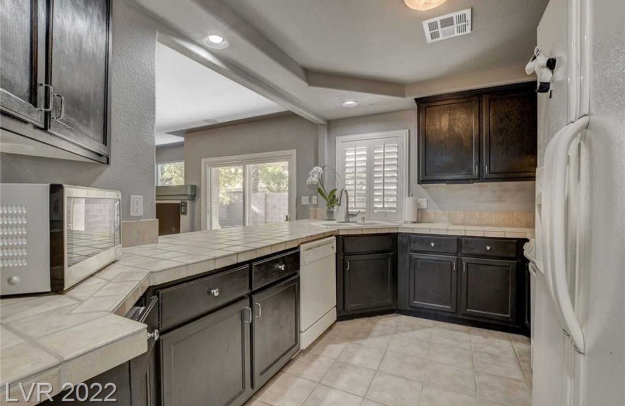 Kitchen with dark cabinets, light countertops, and white appliances; window with shutters.