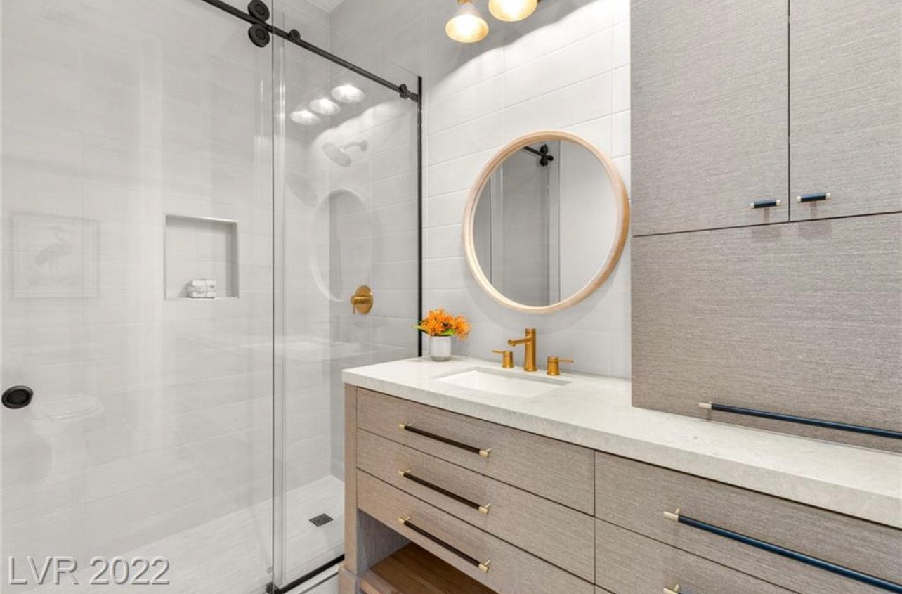 Modern bathroom with a glass shower, vanity, and round mirror. Neutral colors, gold fixtures.