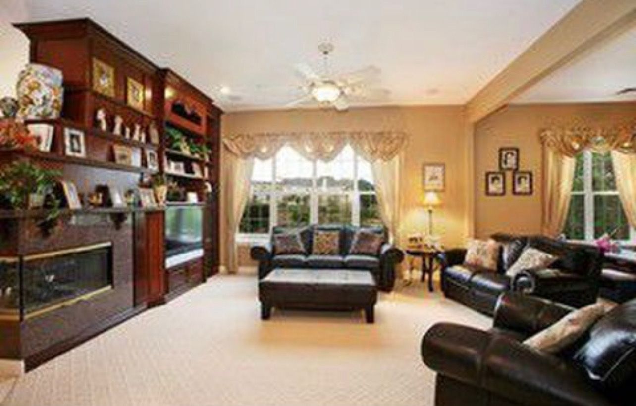 Living room with dark wood built-in shelving and fireplace. Brown leather furniture, beige walls, and large windows.