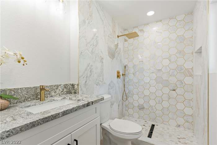 Bathroom with marble walls and hexagon shower tile, gold fixtures, and white vanity.