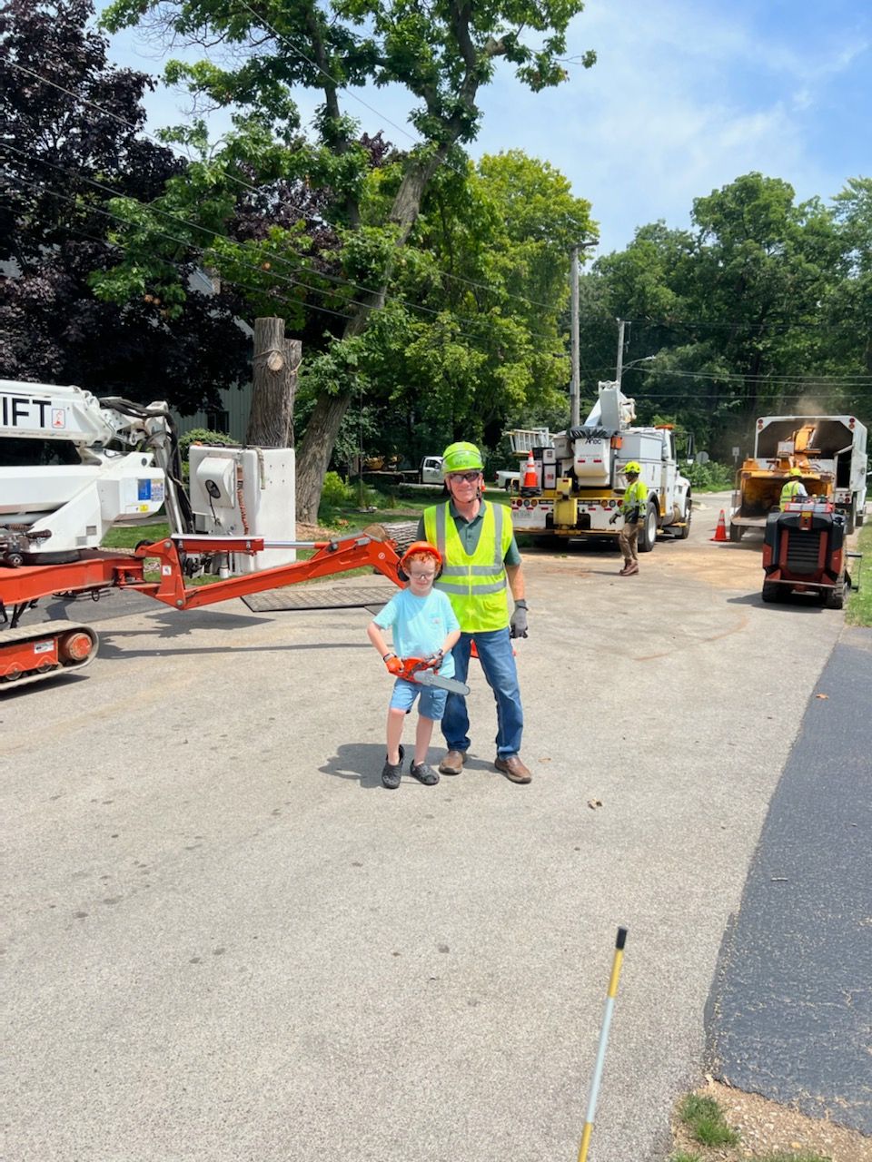 Man and boy stand near work trucks on a sunny day. They wear safety vests and hard hats.