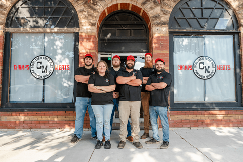 Five people in black shirts pose outside a storefront with arched windows and a red brick facade.