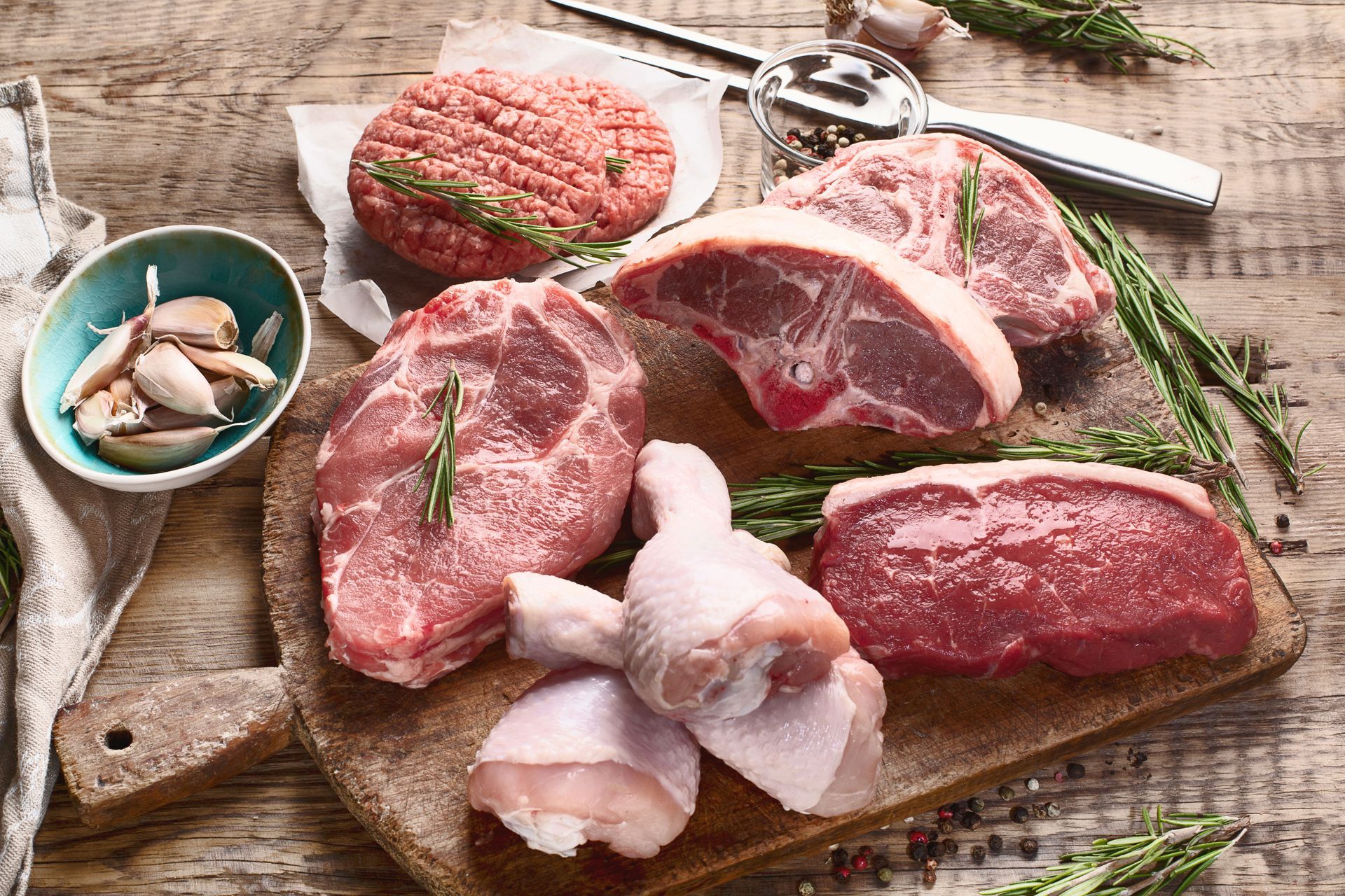 Raw assorted meats and garlic on a wooden cutting board with herbs and kitchen scissors