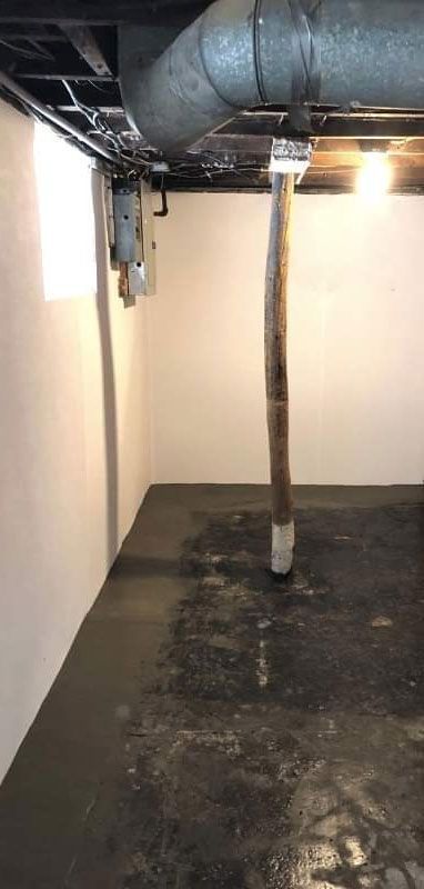 Basement with damp floor and overhead pipes; vertical drainpipe; white walls.