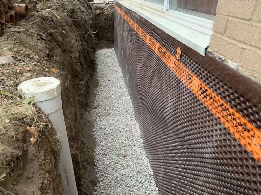 Trench next to a building foundation filled with gravel and a black dimpled drainage membrane.