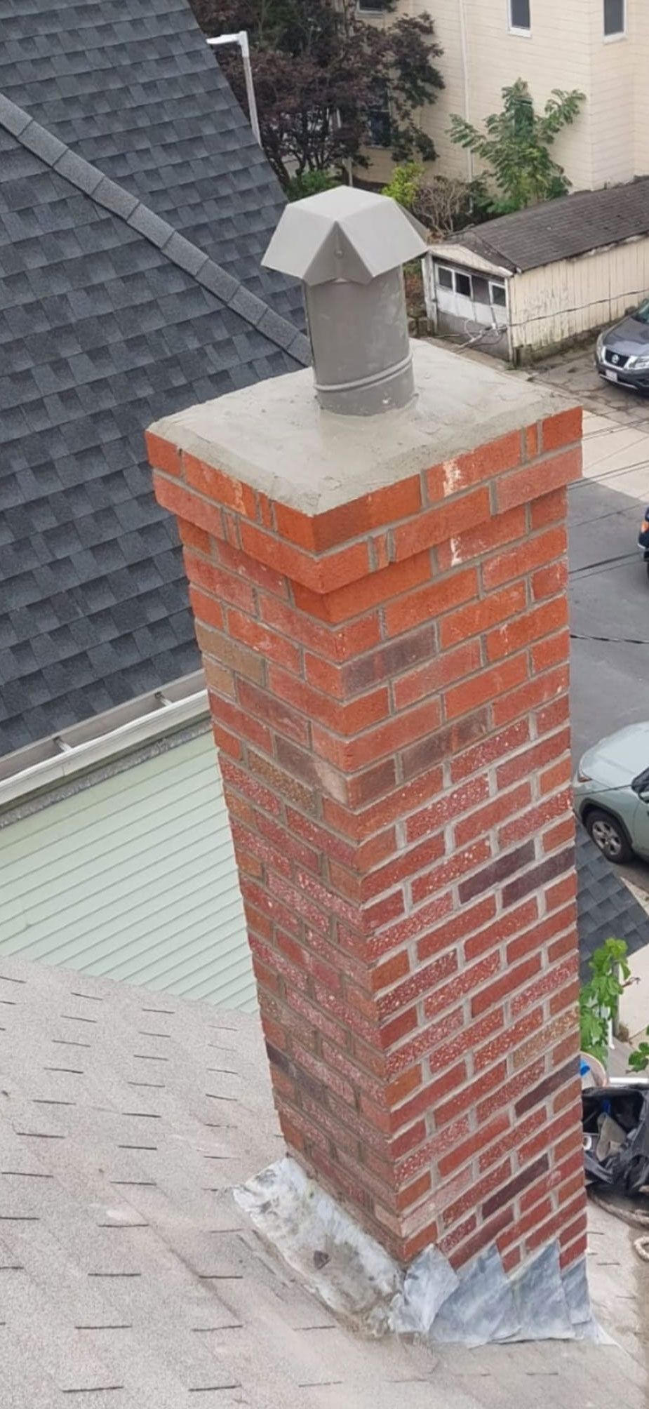 Brick chimney on a gray shingled roof. A metal cap is on top of the chimney.