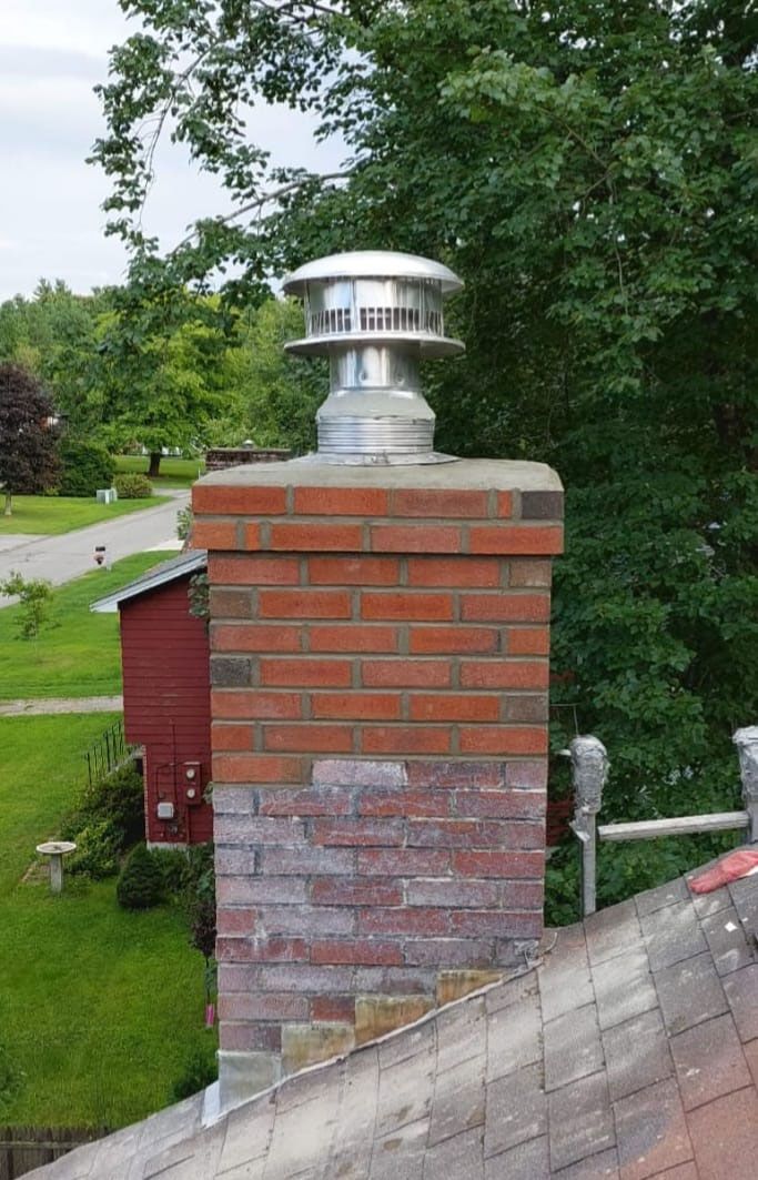 Brick chimney with a metal cap, viewed from a roof, set against trees and a distant red building.