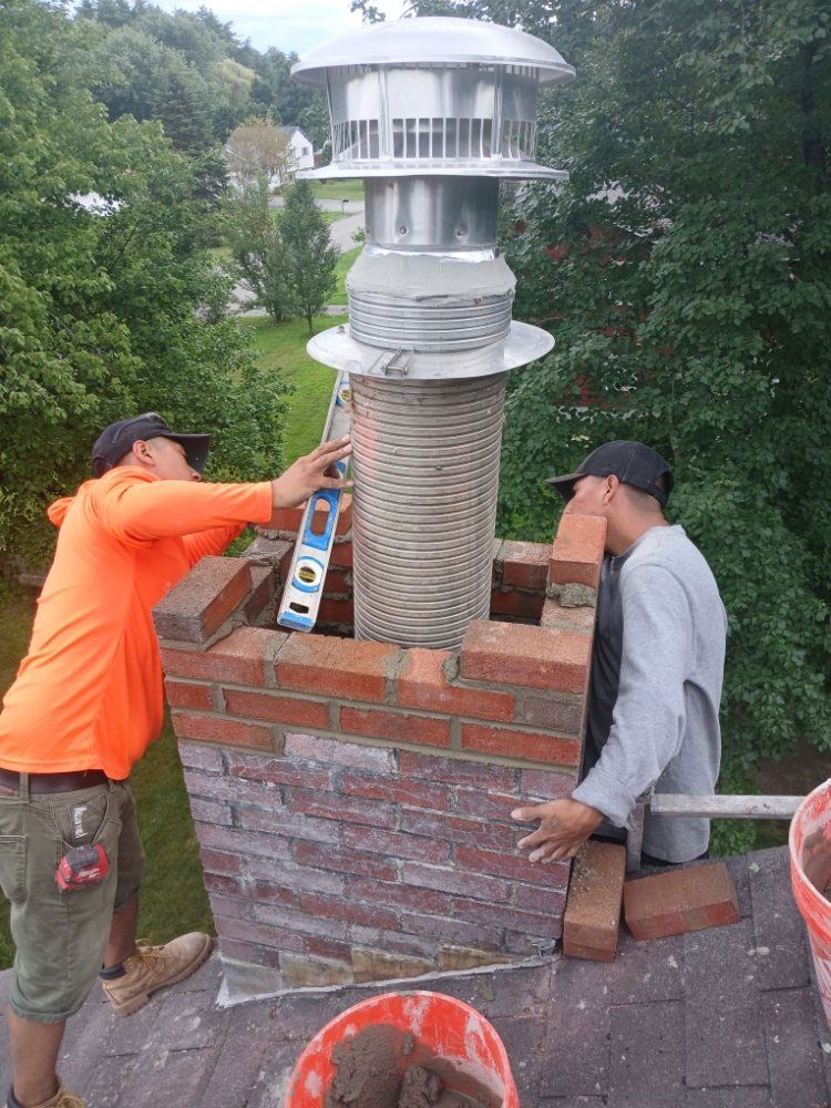 Two men rebuilding a brick chimney on a rooftop; one uses a level, the other assists.