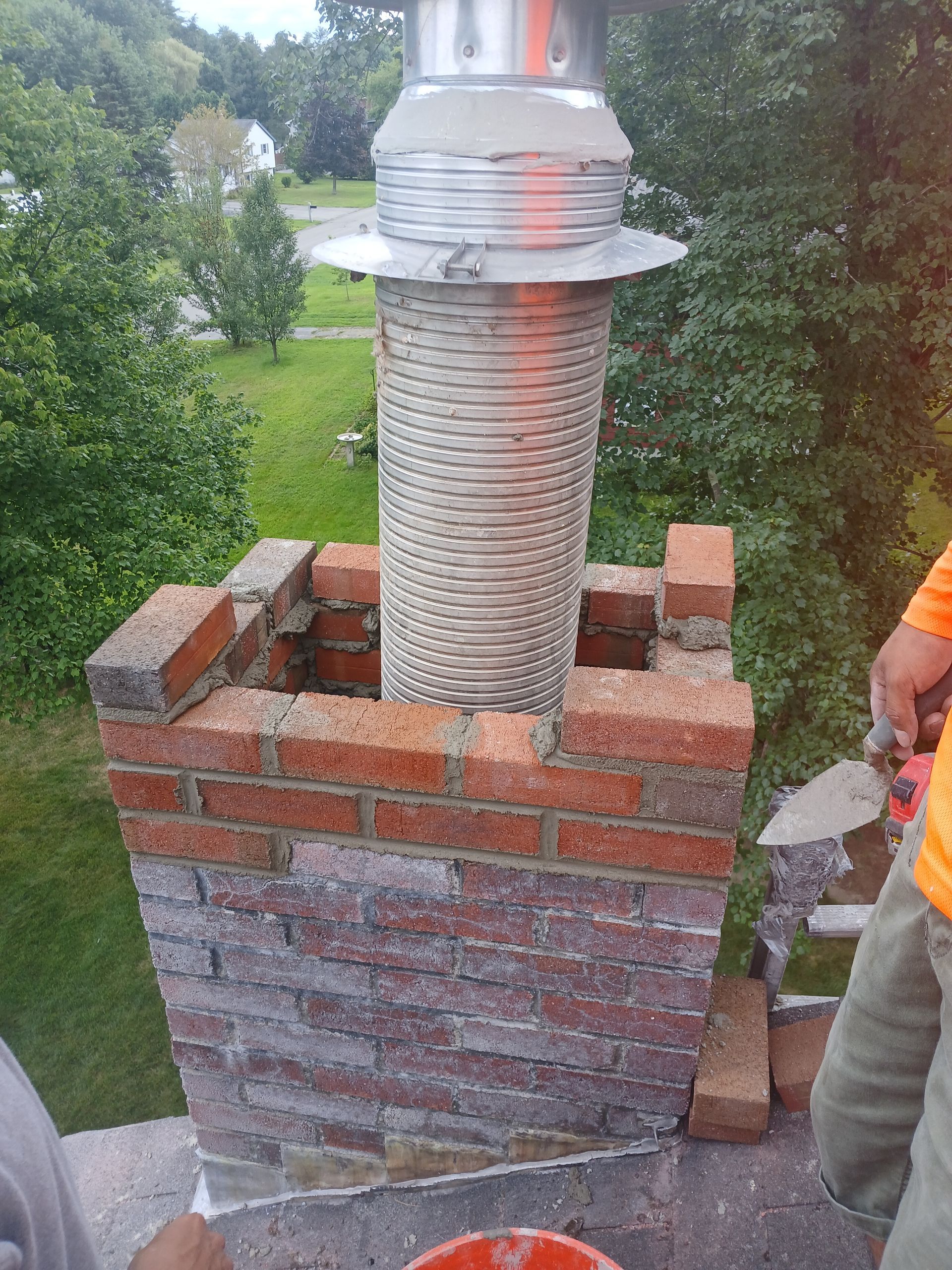Chimney under construction; red brick structure with metal flue, a person's arm is visible, green background.
