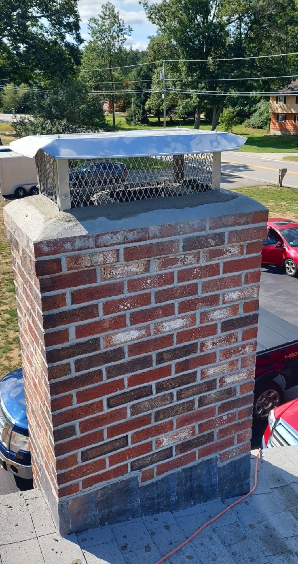 Brick chimney on a rooftop with a metal cap, surrounded by green trees and a blue sky.