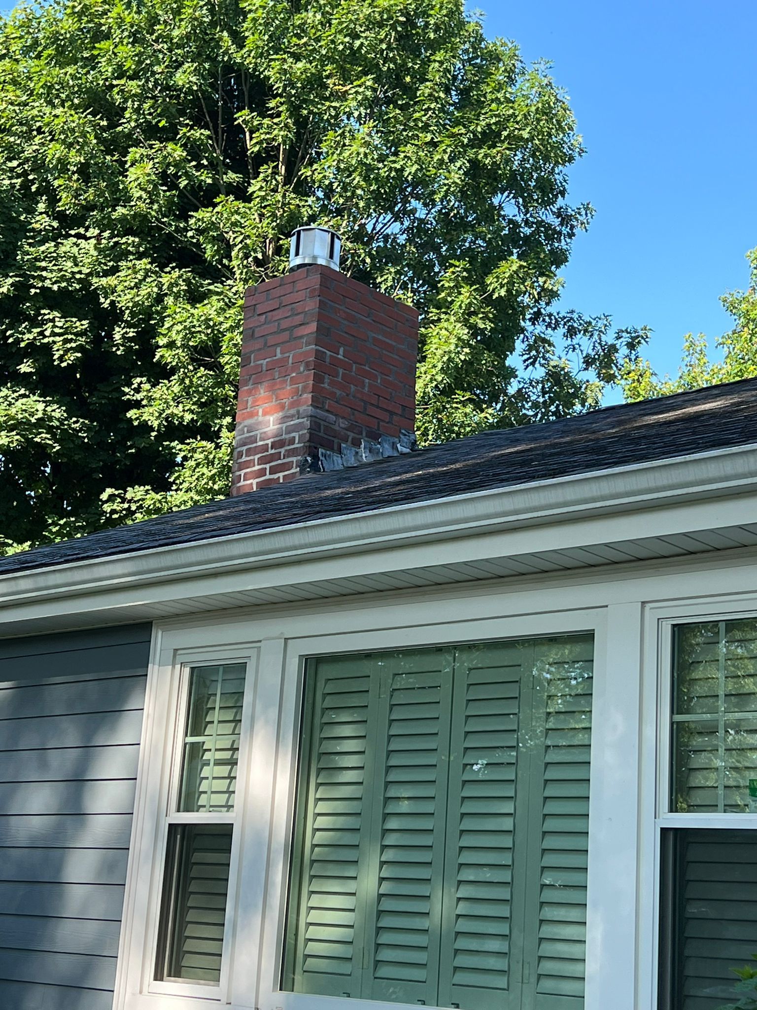 Brick chimney on a roof with green shutters and siding, set against a blue sky and trees.