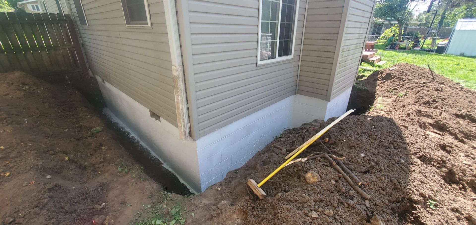 A house with a trench dug around the foundation, and a rake lying on the soil next to it.