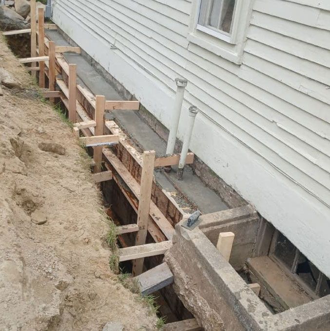 Construction of a concrete foundation along the side of a house; wooden forms and excavated earth visible.