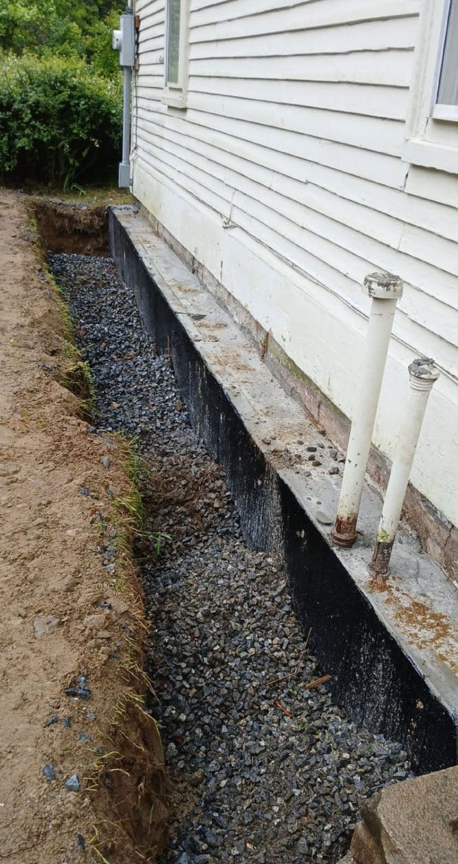 Gravel-filled trench along the white siding of a building. Two pipes protrude.