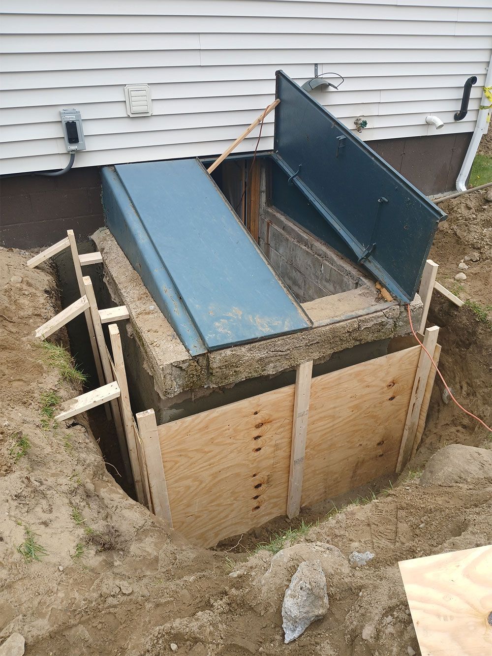 Concrete basement entry with open blue metal doors, surrounded by plywood forms and earth, next to a house.