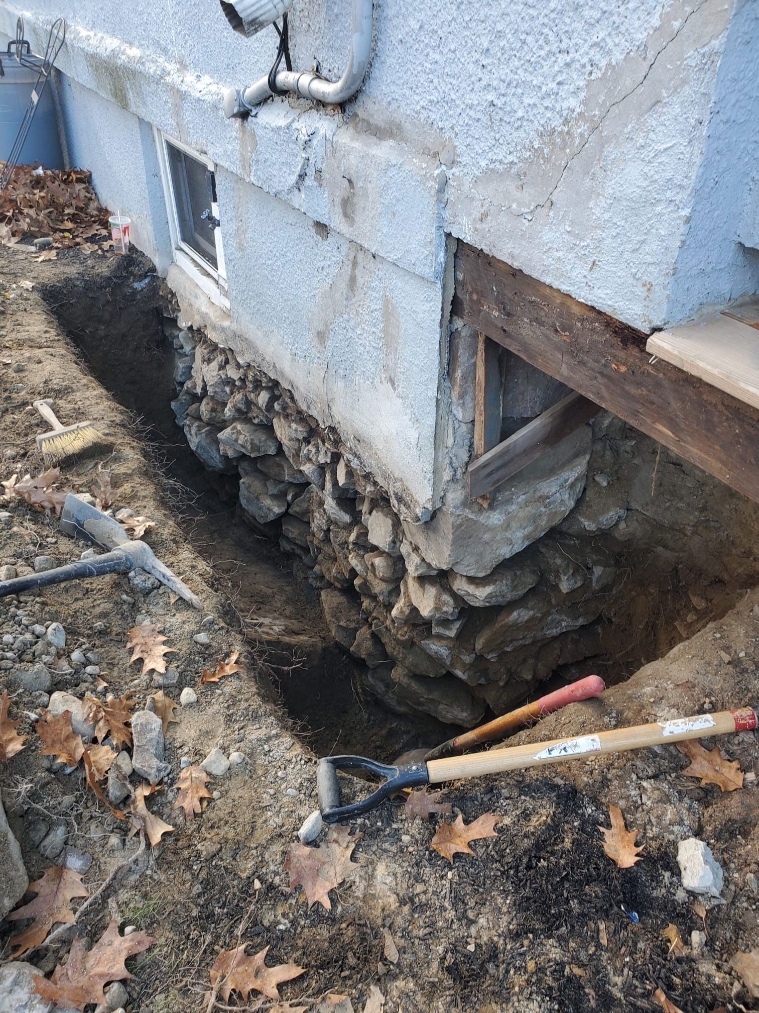 A trench dug along a stone foundation of a building, with tools and fallen leaves nearby.