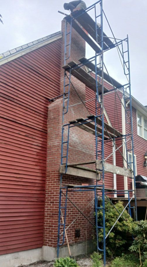 Person working on brick chimney from a blue scaffolding against a red-sided building.