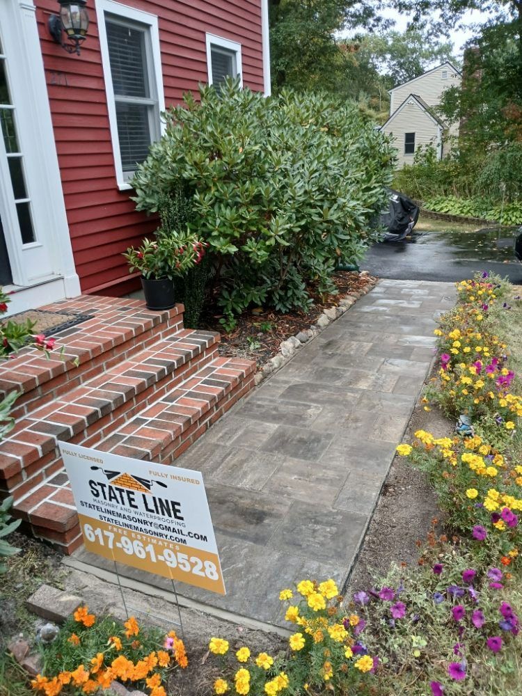 Brick steps, paved walkway, and flower beds lead to a red house; State Line sign visible.